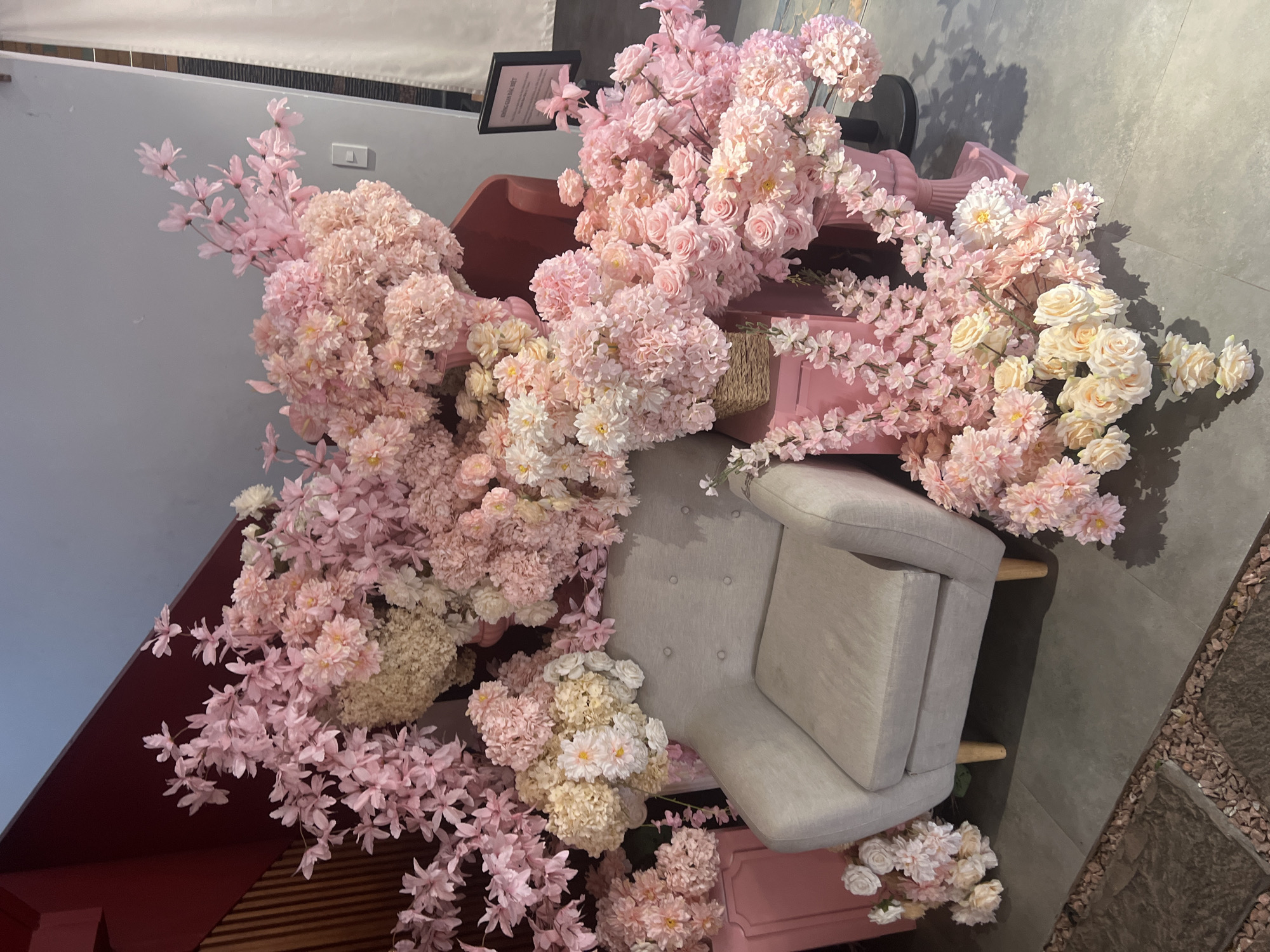 The image shows an indoor scene with a light gray armchair surrounded by an abundance of pink and white flowers. The flowers appear to be arranged around and on top of a pink decorative structure that is behind the chair. The background includes a gray wall and part of a dark red structure. The floor is a mix of dark gray tiles and an area with small brown stones. A small black sign is visible in the background. The location is Quy Nhon Ward, Vietnam. No people are visible in the image.