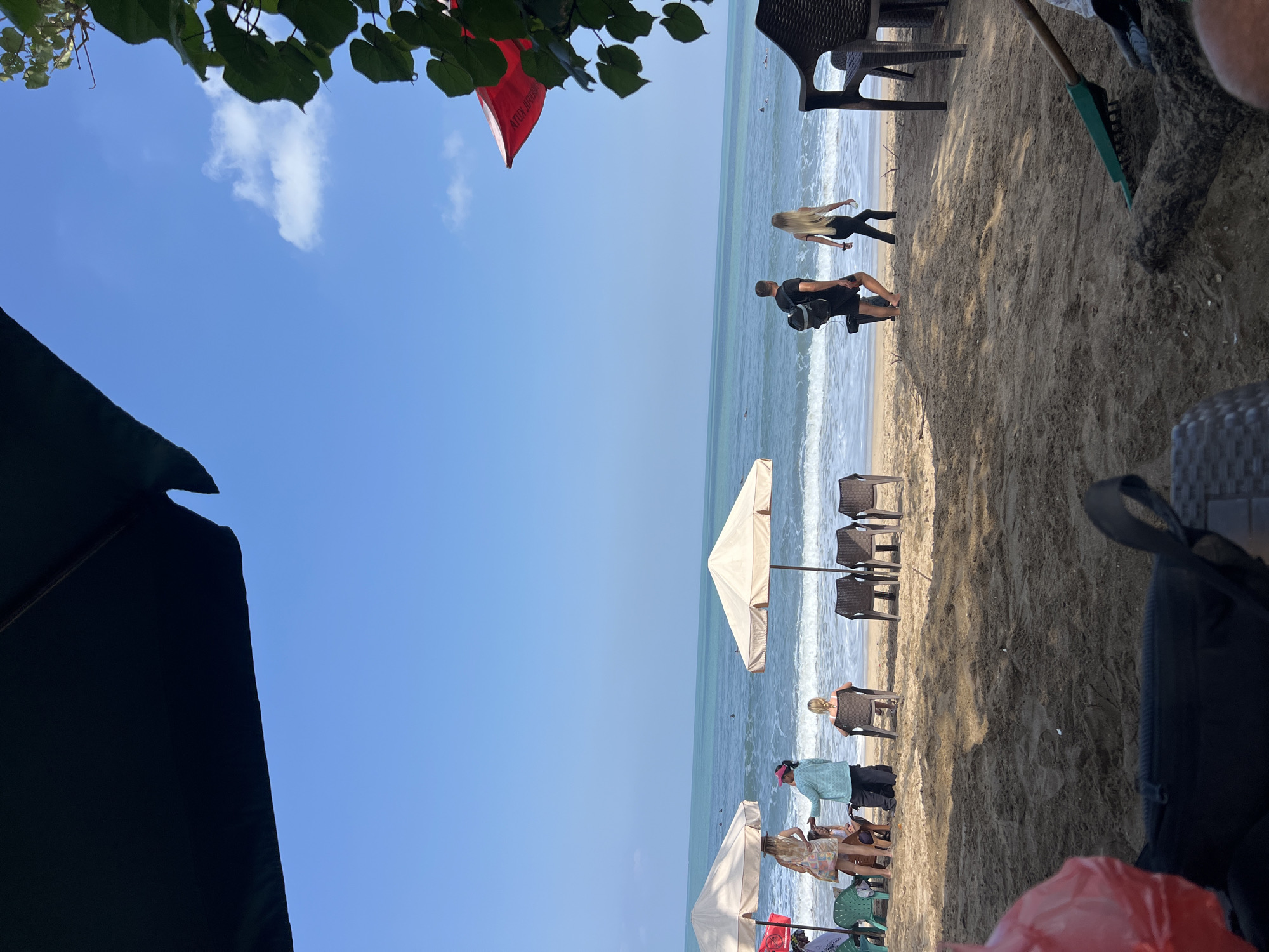 A beach scene on a sunny day in Kuta, Indonesia, shows several people relaxing and walking by the ocean. The foreground is dominated by sand and shadows from overhead umbrellas, with some beach chairs and a rake visible. Further back, people are seated under beach umbrellas, some engaging in conversation. A couple walks along the shore towards the water, while others are seen in the distance in the turquoise ocean, some appearing to be surfing. The sky is clear blue with a few wispy clouds.
