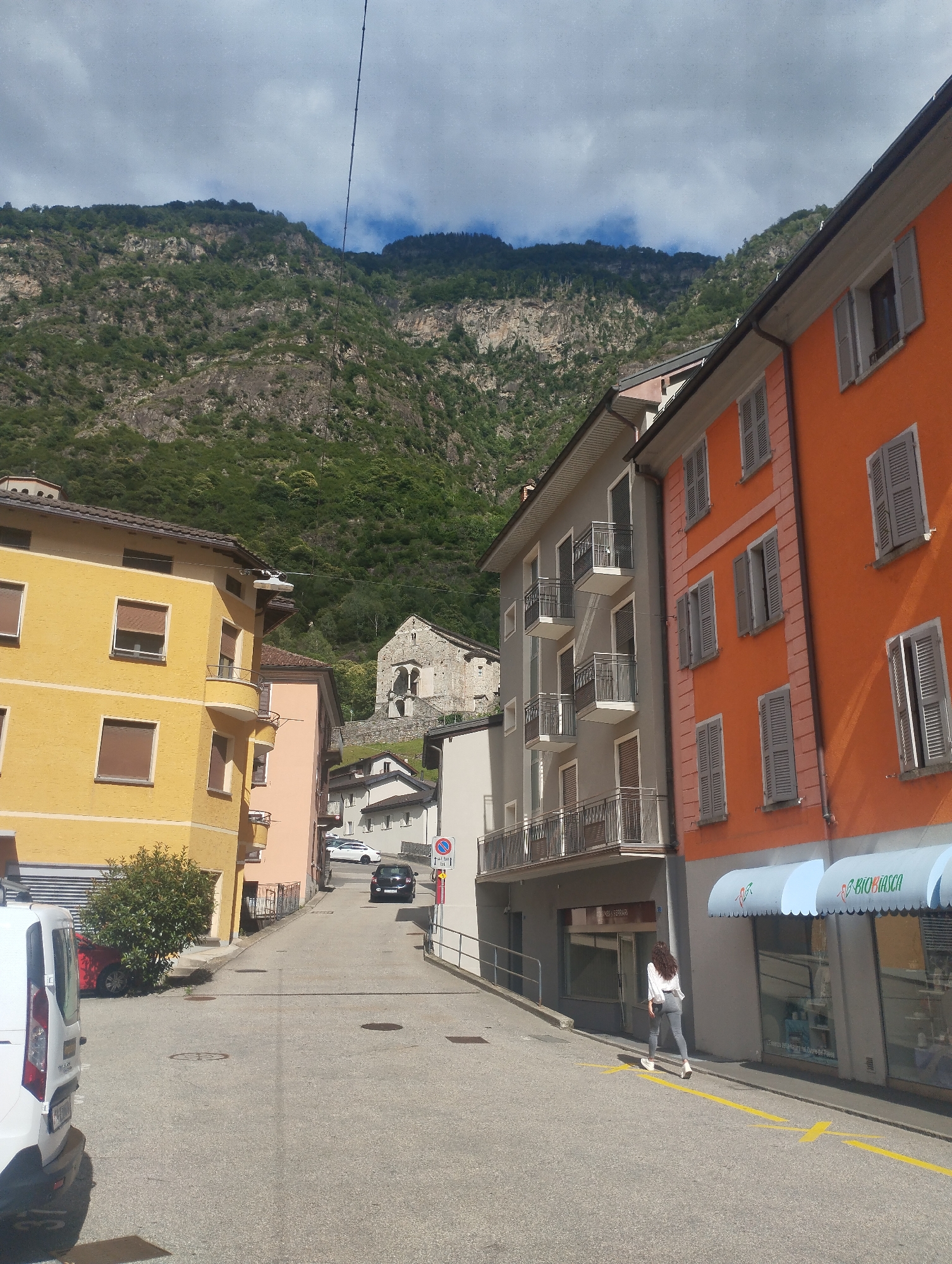 A street scene in a European town is depicted, nestled at the base of a steep, verdant mountain. Buildings with colorful facades, primarily orange and yellow, line the street, some featuring balconies and shutters. A woman in a white shirt and gray pants walks away from the viewer on the right side of the street, following a yellow line painted on the asphalt. A white van is parked on the left side of the street, and a dark car is driving away from the viewer further down the road. In the background, a stone structure, possibly a church or ruin, is visible on a hillside. The sky is partly cloudy, and the overall atmosphere suggests a sunny day in a mountainous region.