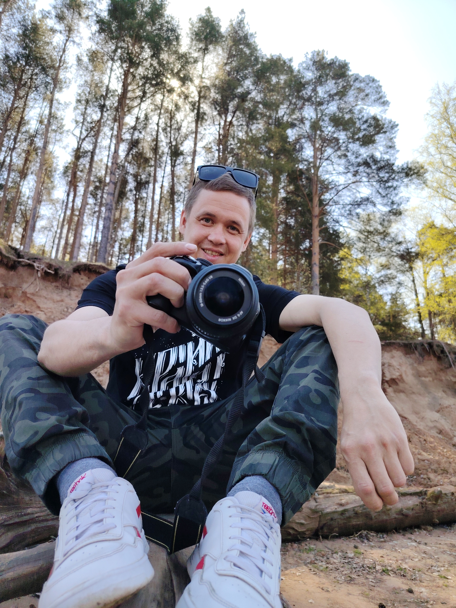 A medium shot captures a smiling Caucasian man holding a black camera. He is sitting cross-legged on a log with his white sneakers in the foreground. He is wearing camouflage pants, a black t-shirt with a white design, and sunglasses are perched on his head. The scene is set in a wooded area, likely in Chaikovsky, Russia. The ground is sandy and light brown. Tall trees with light green foliage rise into the sky behind him. Some sunlight is visible through the trees. The man is holding the camera up as if taking a picture.