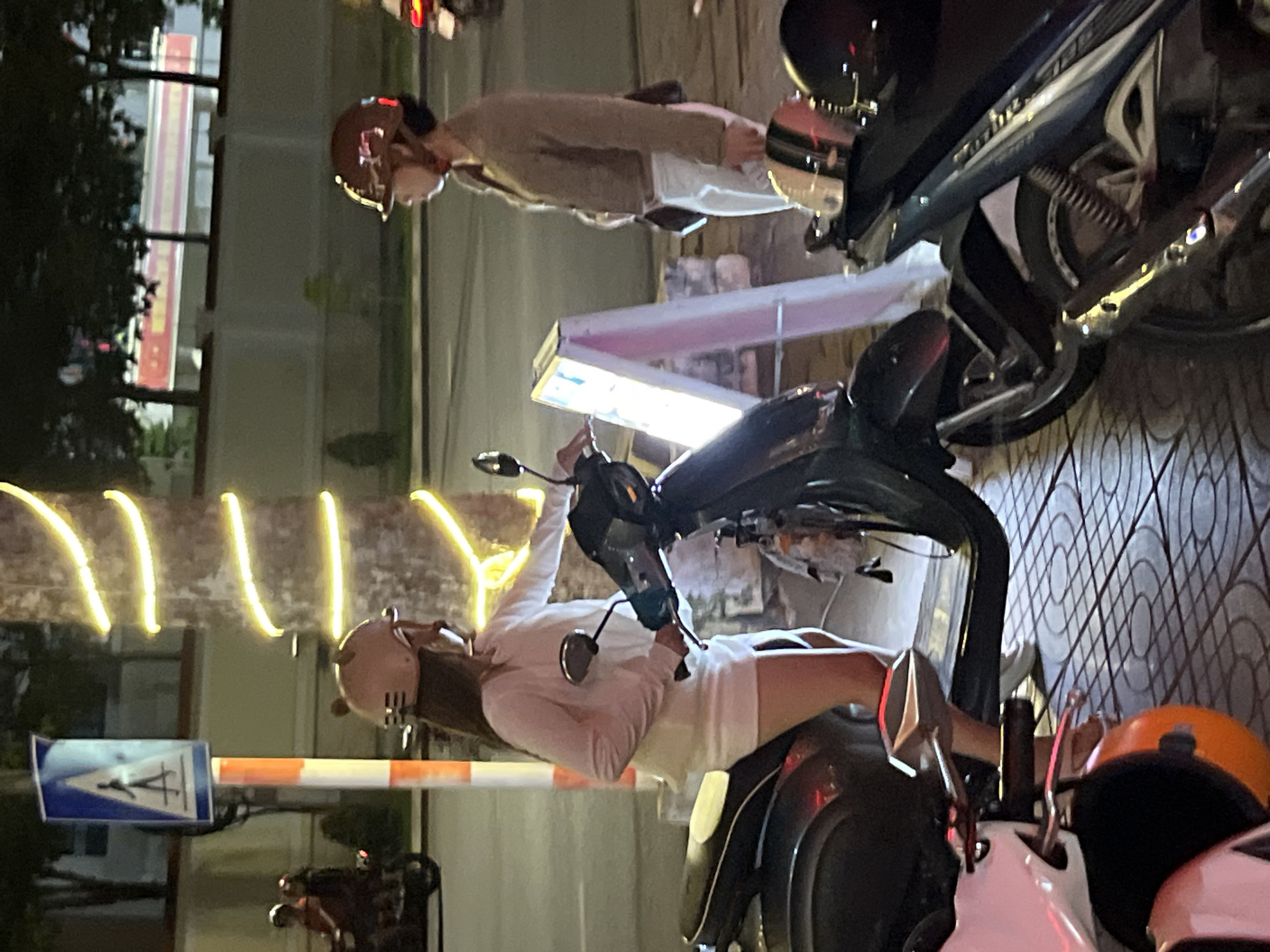 In Phu Thuy Ward, Vietnam, two women are seen near motorcycles at night. One woman, wearing a light-colored long-sleeved shirt and a white skirt, stands with her hand on the handlebars of a dark-colored scooter. She has a light brown helmet on and appears to be interacting with the scooter. Next to her, another woman wearing a brown jacket and white pants stands facing away from the camera. She also wears a helmet. A lighted sign is positioned between the two women and the scooters. In the background, a traffic sign indicating a pedestrian crossing is visible, along with a tree wrapped in yellow lights. Several motorcycles are parked around the area.