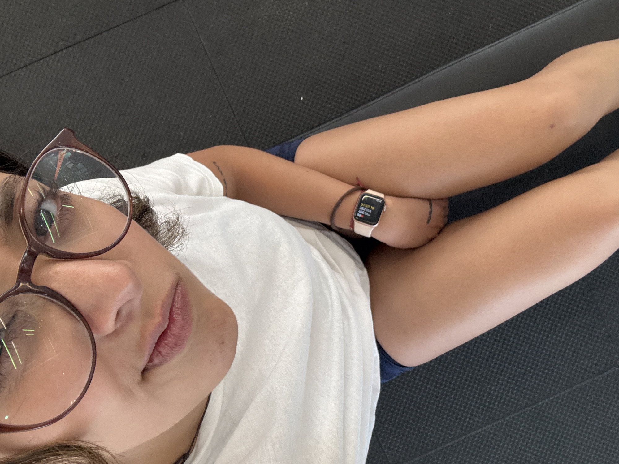 A person wearing glasses, a white t-shirt, and an Apple watch is sitting on a dark textured surface, possibly a bench or mat. The person's legs are crossed. The location may be San Andrés Cholula, Mexico, but there is no discernible evidence to support this.