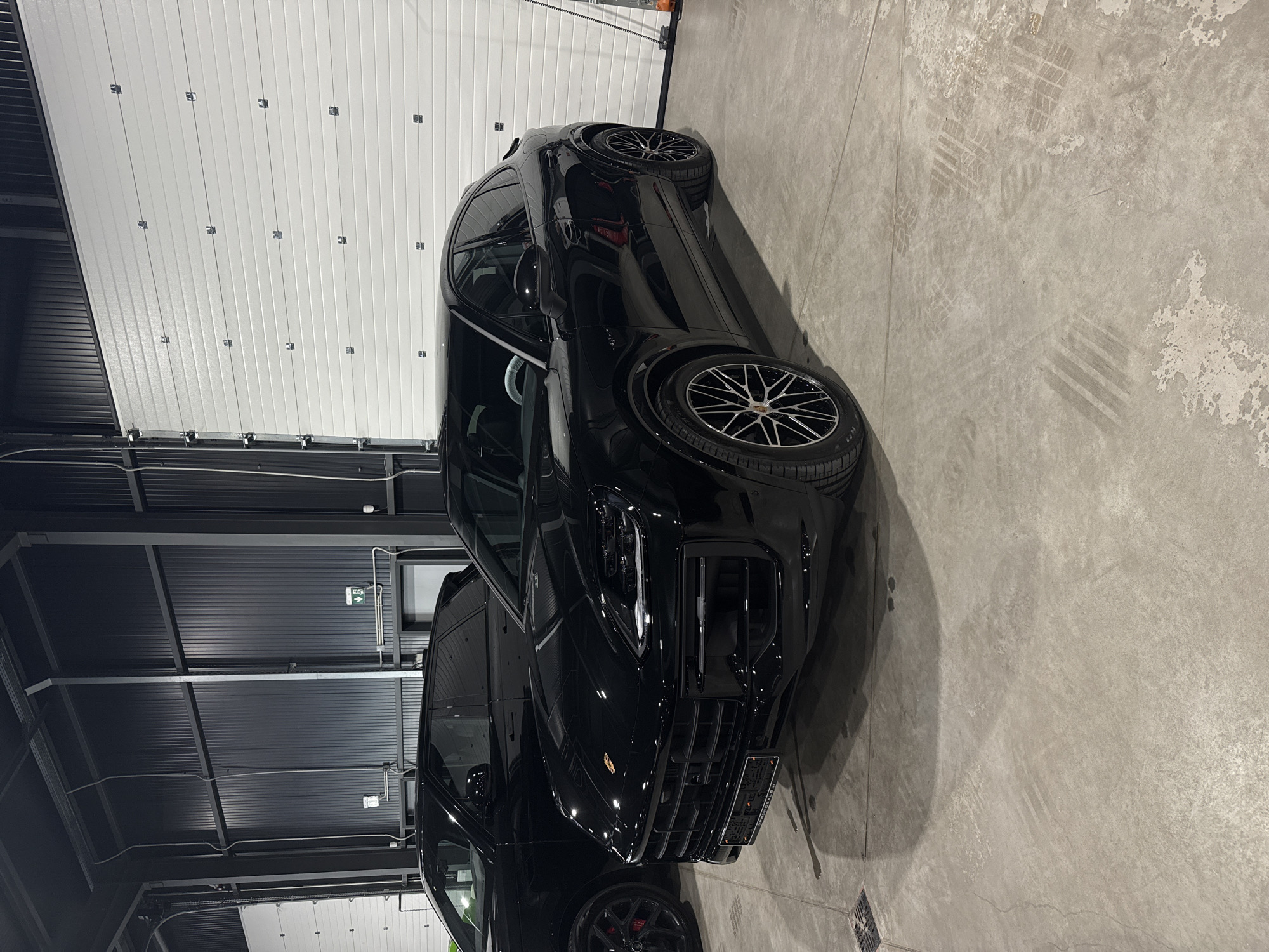 The image displays a highly polished, black Porsche SUV or crossover, resembling a Cayenne Coupe, parked within an indoor industrial setting, likely a garage, workshop, or showroom. The vehicle's immaculate condition suggests it has been recently cleaned or detailed. It features multi-spoke wheels with a silver and black design, and a prominent red brake caliper is visible on the front left wheel.
The surrounding environment includes a light grey concrete floor with visible tire tracks and areas of surface wear. On the left side of the frame, white sectional garage doors are present. The upper structure of the facility, including the ceiling, is composed of dark grey corrugated metal panels, from which exposed pipes, electrical conduits, and artificial light fixtures hang. A green emergency exit sign with a white arrow pointing to the right is mounted on the ceiling.
The scene is static, with no individuals present. A European-style license plate on the front of the car partially displays "B 8" followed by obscured characters, with "BG" clearly visible below, confirming its location in Bulgaria, consistent with the provided context of Plovdiv. Text indicating tire specifications, such as "P ZERO N1," is legible on the sidewall of the front tire. The image portrays a clean, modern vehicle against a functional, utilitarian backdrop.