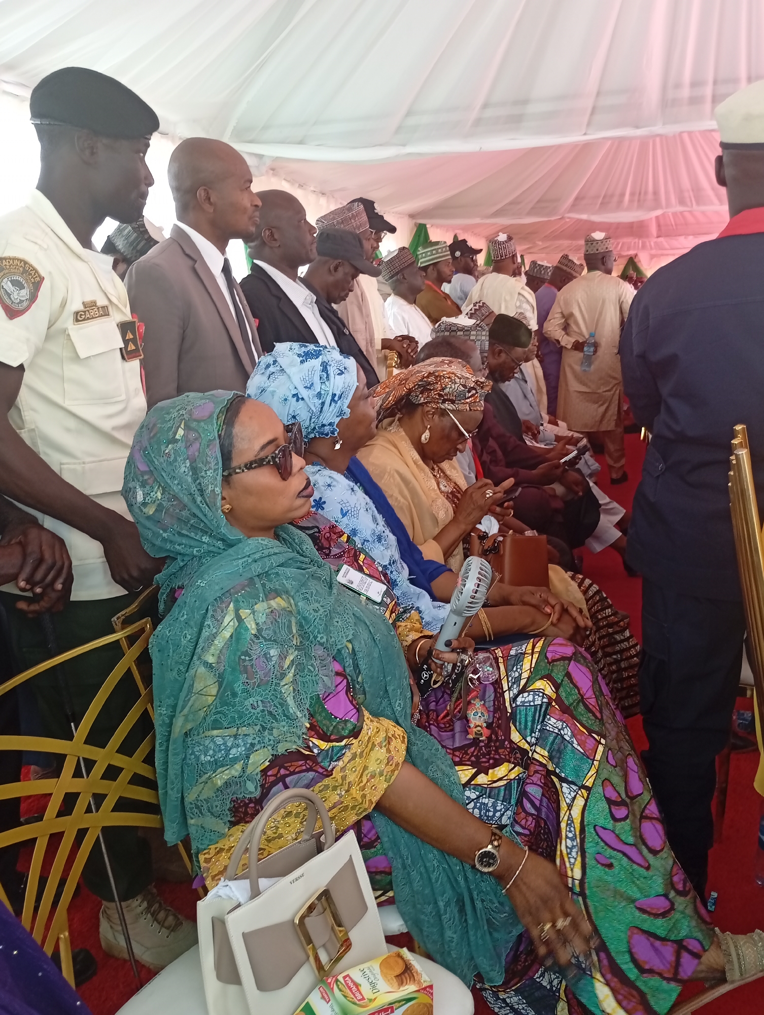 The image depicts a large gathering of people, both men and women, attending an event under a large white canopy tent, likely outdoors during the day in Unguwan Sanusi, Nigeria.
In the left foreground, a man in a beige uniform with a black beret stands, looking to the right. His uniform bears a patch with "KADUNA STATE" and an emblem, along with the name "GARBA". Adjacent to him are other men dressed in suits and traditional African attire, including various caps.
Seated in the foreground are several women. Prominently featured is a woman on the left wearing a green patterned hijab and a vibrant multi-colored dress with patterns in green, purple, yellow, and blue. She is also wearing sunglasses, a wristwatch, and rings. Next to her, another woman wears a blue patterned headscarf. A third woman in a gold-colored head wrap and top is visible further back, holding a small white portable fan. Many attendees, both men and women in the background, are observed using or looking at their mobile phones.
The setting includes golden-framed chairs and a red carpet, suggesting a formal or semi-formal occasion. On a surface beside the woman in the green hijab, a light-colored handbag with the brand name "VERSSI" is visible, along with a package of "Britannia Digestive" biscuits. The presence of the portable fan indicates warm weather.