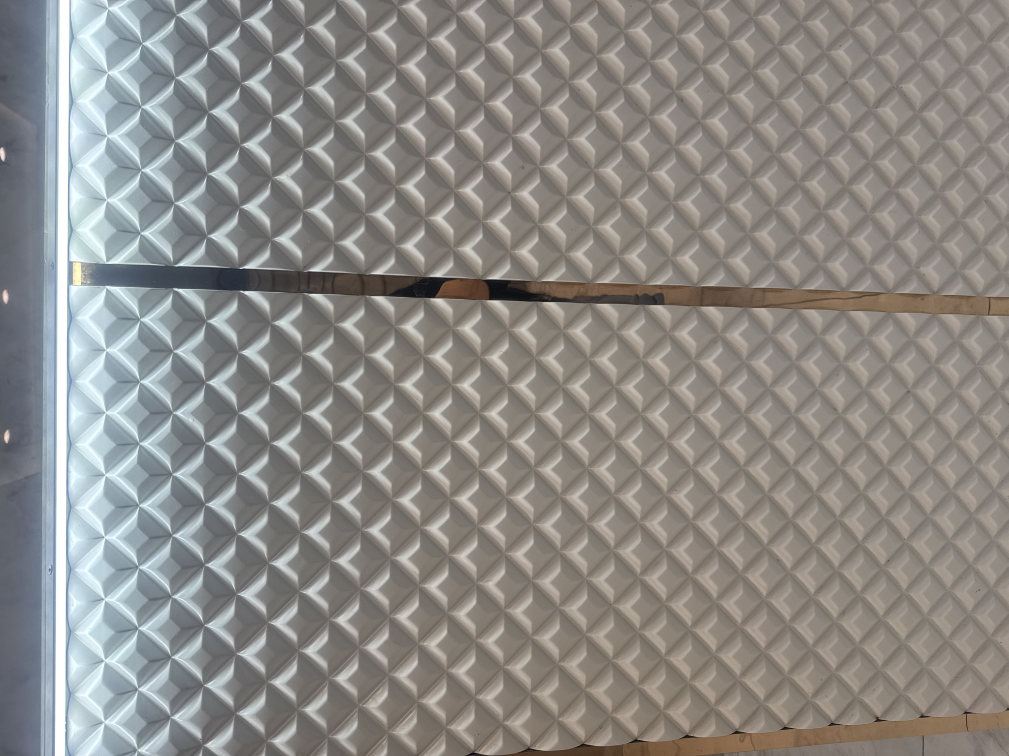 The image depicts a textured white wall panel with a repeating geometric pattern of raised squares, segmented by two horizontal gold-colored strips. The panel is installed in what appears to be an interior space. The left edge of the panel is illuminated by a vertical light source. The image was taken in the city of Yerevan, Armenia.