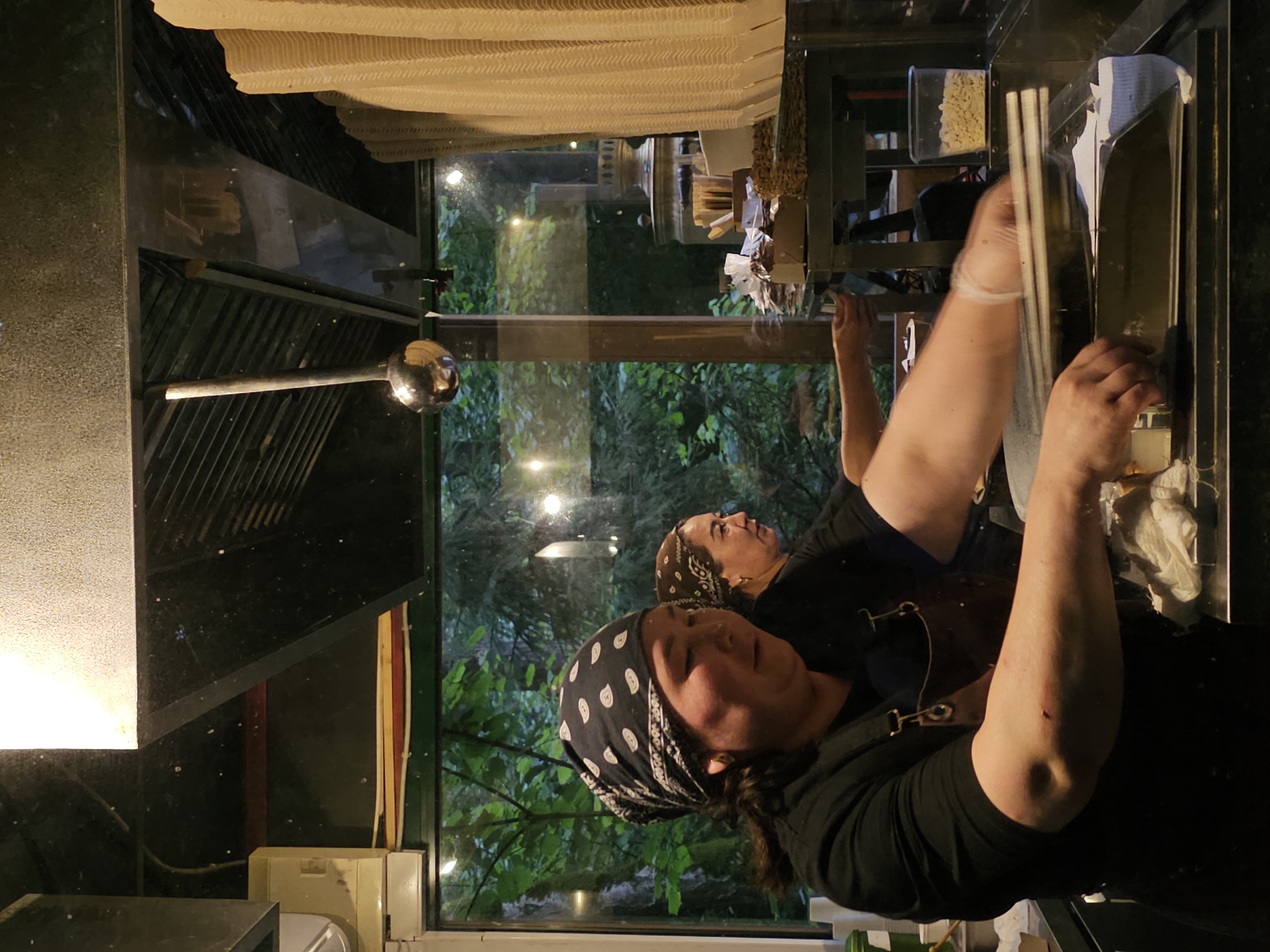 In a food service establishment in Nemchinovo, Russia, two women are engaged in food preparation behind a counter. The woman in the foreground, wearing a black shirt and apron, is focused on her task, with her hands blurred by motion as she handles what appear to be chopsticks over a metal tray. She has a bandana with a white paisley pattern on her head. Behind her, another woman, also wearing a bandana, is visible in profile, her gaze directed to her right. The environment suggests a commercial kitchen setting, with various utensils and containers present. The background, visible through a window, shows a lush, green outdoor environment with trees, hinting at the establishment's location.