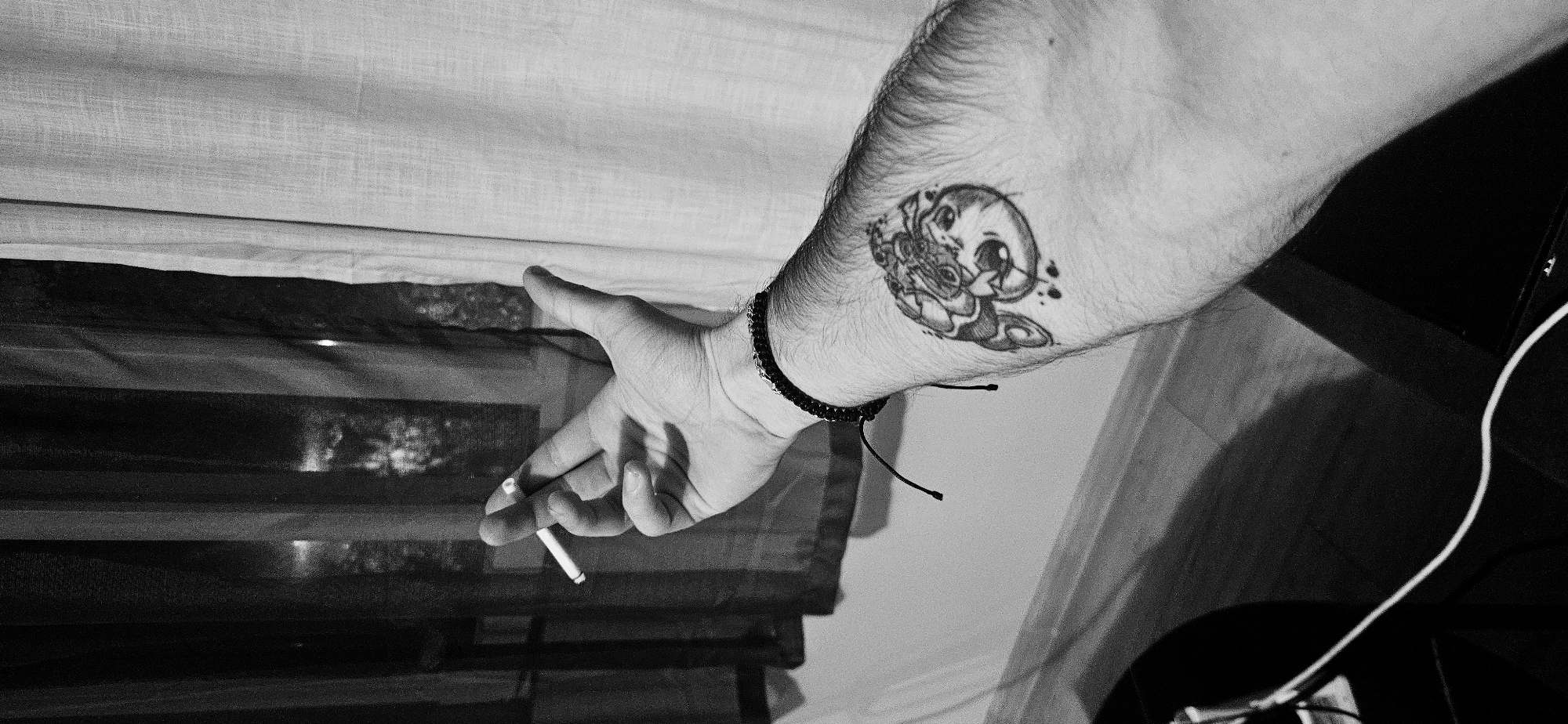 A black and white close-up photograph depicts a person's arm, hand, and a portion of their torso, with a cigarette held between their fingers. The setting appears to be indoors.
The person's arm, adorned with a tattoo, is positioned diagonally across the frame. A beaded bracelet is visible on the wrist. The hand holds a lit cigarette between the index and middle fingers.
In the background, a semi-transparent curtain is partially visible. Below the curtain is a dark, horizontal, barred structure. To the right of the arm, a white cable is visible, and a portion of a dark object.
The scene lacks specific landmarks to pinpoint its location within Plovdiv, Bulgaria. Without additional visual or contextual information, such as recognizable street signs, buildings, or specific interior details, it is impossible to determine the exact location within the city.