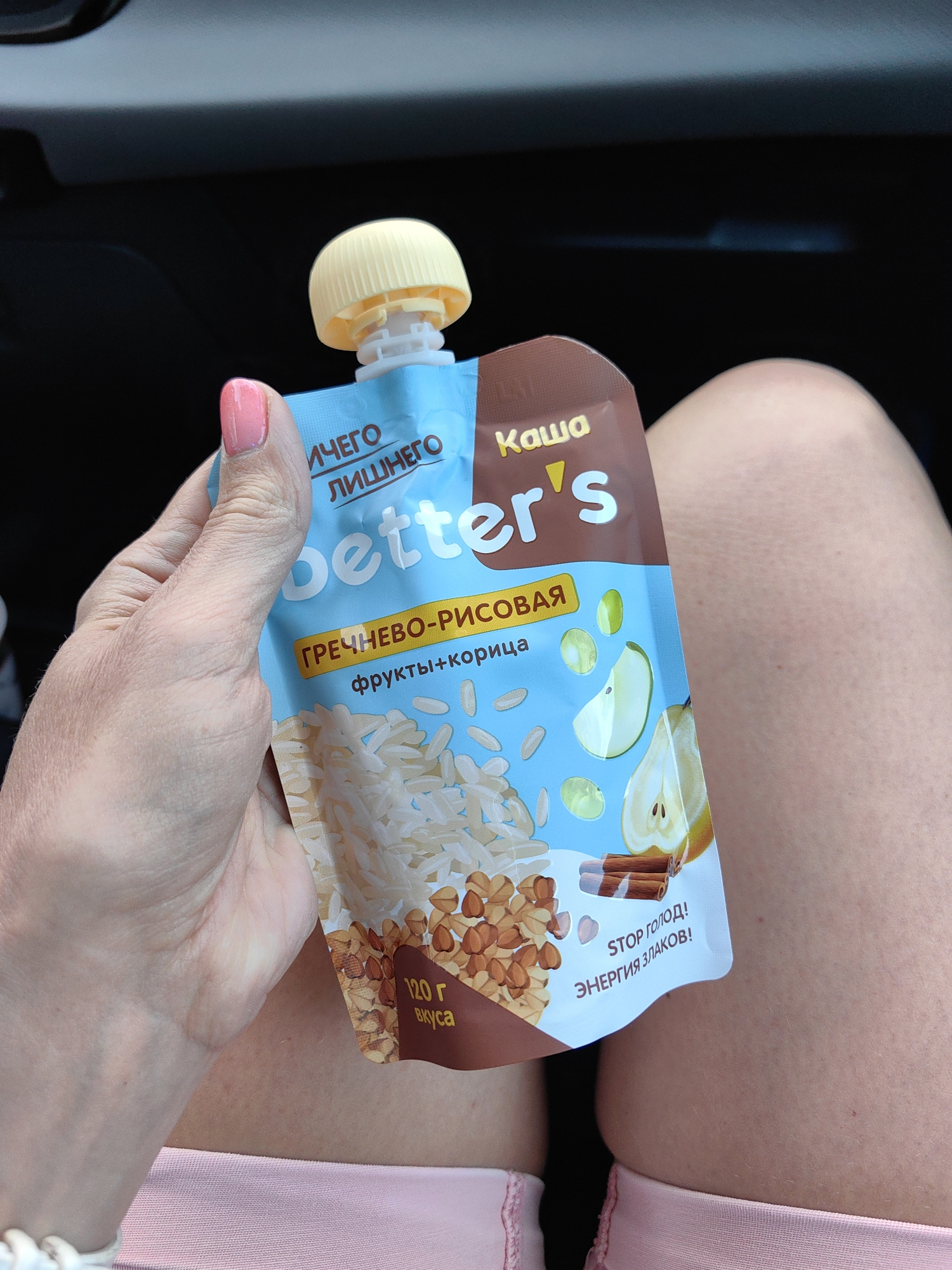 An overhead shot shows a person's hand holding a pouch of baby food. The pouch is light blue with illustrations of rice, buckwheat, pear slices, and cinnamon sticks. The brand name "Better's" is written in large white letters. Text on the pouch reads "ГРЕЧНЕВО-РИСОВАЯ" (buckwheat-rice), "фрукты+корица" (fruit+cinnamon), "120 г вкуса" (120 g of taste), and "STOP ГОЛОД! ЭНЕРГИЯ ЗЛАКОВ!" (Stop hunger! Energy of grains!). The pouch has a yellow cap. The background is dark, suggesting an indoor or car setting. The image is cropped and does not provide enough information to determine the exact location within Крывасельскі сельскі Савет, Belarus.