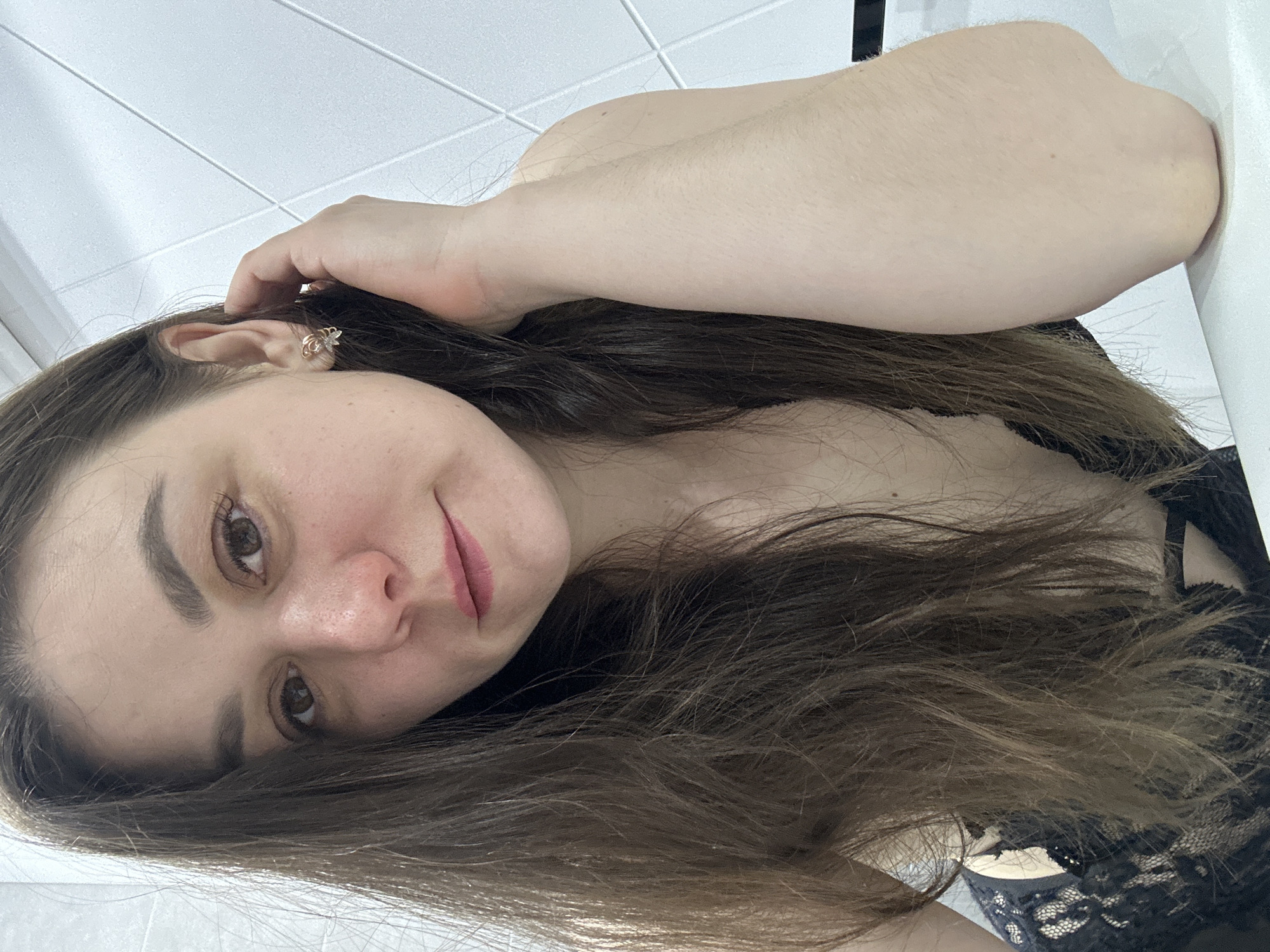 A young woman is captured in a close-up, top-down portrait, likely a selfie. She is lying down, with her head angled towards the camera, displaying a gentle, relaxed expression and a subtle smile. Her long, dark brown hair is spread around her head and upper body. Her clear skin, dark eyebrows, and light brown eyes are prominent. A small, decorative earring, featuring a white stone or pearl and gold-toned metal, is visible in her right ear. Her right arm is bent, with her hand near her head. She is wearing a black lace garment, partially visible on her chest and shoulder. The background suggests an indoor setting, with a white ceiling featuring a grid pattern discernible behind her. Her arm appears to rest against a white surface, possibly a wall or furniture. No text is visible in the image. The scene is set in Chaikovsky, Russia.