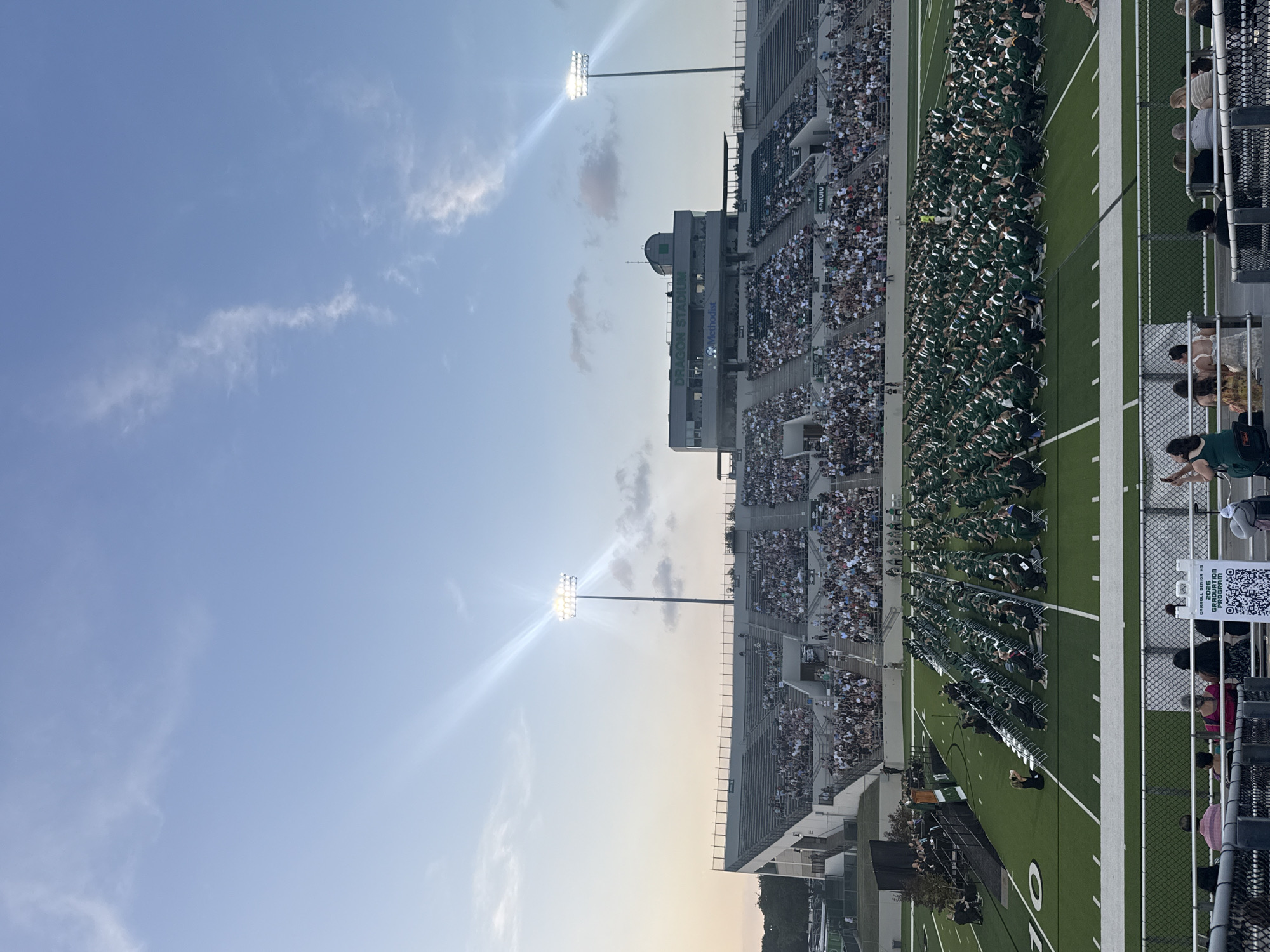 The image captures a graduation ceremony at Dragon Stadium in Southlake, Texas, United States of America. Graduates wearing green gowns and white caps are seated in rows on the green football field. The stands are filled with spectators. In the upper left, the sky is blue with some clouds and bright stadium lights. The structure bearing the name "Dragon Stadium" is visible, along with a Methodist logo. A fence and some people are in the extreme right foreground. A QR code is affixed to the fence.