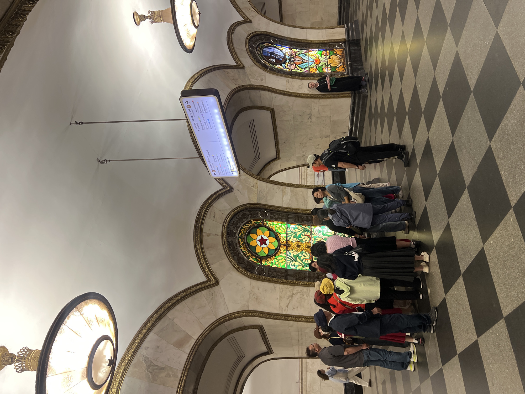 Here is a detailed description of the image:
**Overall Impression:**
The image appears to be taken inside a metro station or an elaborate public building. The interior has arched architecture with stained-glass windows and a checkerboard tiled floor. There is a group of people gathered, seemingly looking up at something of interest.
**Environment:**
The architectural design features archways with decorative golden trim. Stained-glass windows are visible behind the archways. A suspended information board hangs from the ceiling.
**People:**
A group of people is gathered, their attention directed upwards, likely towards the architecture or the stained-glass windows. They are diverse in appearance and attire, wearing casual clothing.
**Objects:**
- Suspended Information Board: A digital information board displaying text in Russian, likely related to metro station names.
- Lighting Fixtures: There are prominent circular lighting fixtures hanging from the ceiling.
- Stained Glass Windows: The stained-glass windows feature intricate designs and are illuminated.
- Checkerboard Floor: The floor is covered with a distinctive black and white checkerboard tile pattern.
**Actions and Interactions:**
The people are standing and looking up. The digital board has names of locations in it, and therefore the interaction is with the location.
**Location:**
The text on the digital board is written in Russian. The names of the metro stops are: Prospect Mira, Novoslobodskaya, Belorusskaya, Krasnopresnenskaya, and others. The place is most likely in Moscow, Russia.