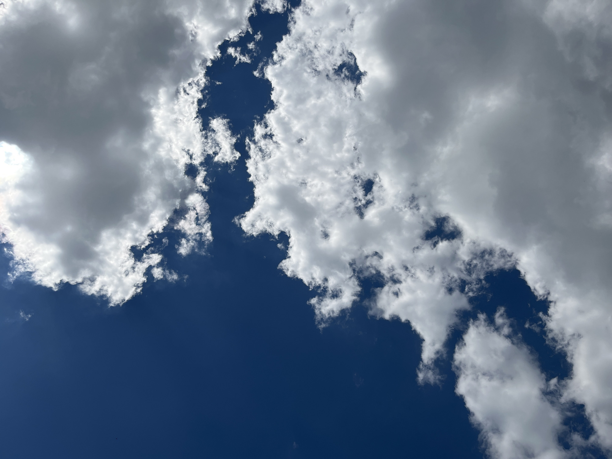 The image shows a sky with scattered clouds and deep blue patches of clear sky. The clouds vary in density and texture, with some appearing bright white and fluffy, while others are darker and more overcast. The sun is visible, creating a bright, almost blown-out area in the upper right portion of the frame, suggesting it is daytime. There are no people, animals, buildings, or other man-made objects visible in the image. The perspective is from below, looking upwards. The image is taken outdoors. The location is Bilenke, Ukraine.
