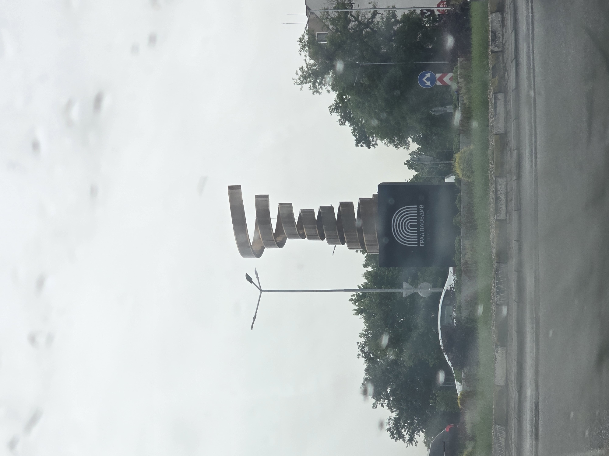 The image depicts an outdoor scene, possibly taken from inside a vehicle due to the presence of raindrops on the glass. The focal point is a modern sculpture constructed from metal, appearing as a series of interconnected, open-ended arches. Beside the sculpture is a dark rectangular sign with vertical stripes, which also displays the text "ГРАД ПЛОВДИВ," written in Cyrillic. This sign suggests that the sculpture is located in Plovdiv, Bulgaria. In the background, there are trees, a traffic sign indicating a sharp turn, and a building. A street and sidewalk are visible along the right edge. Overall, the weather appears overcast.