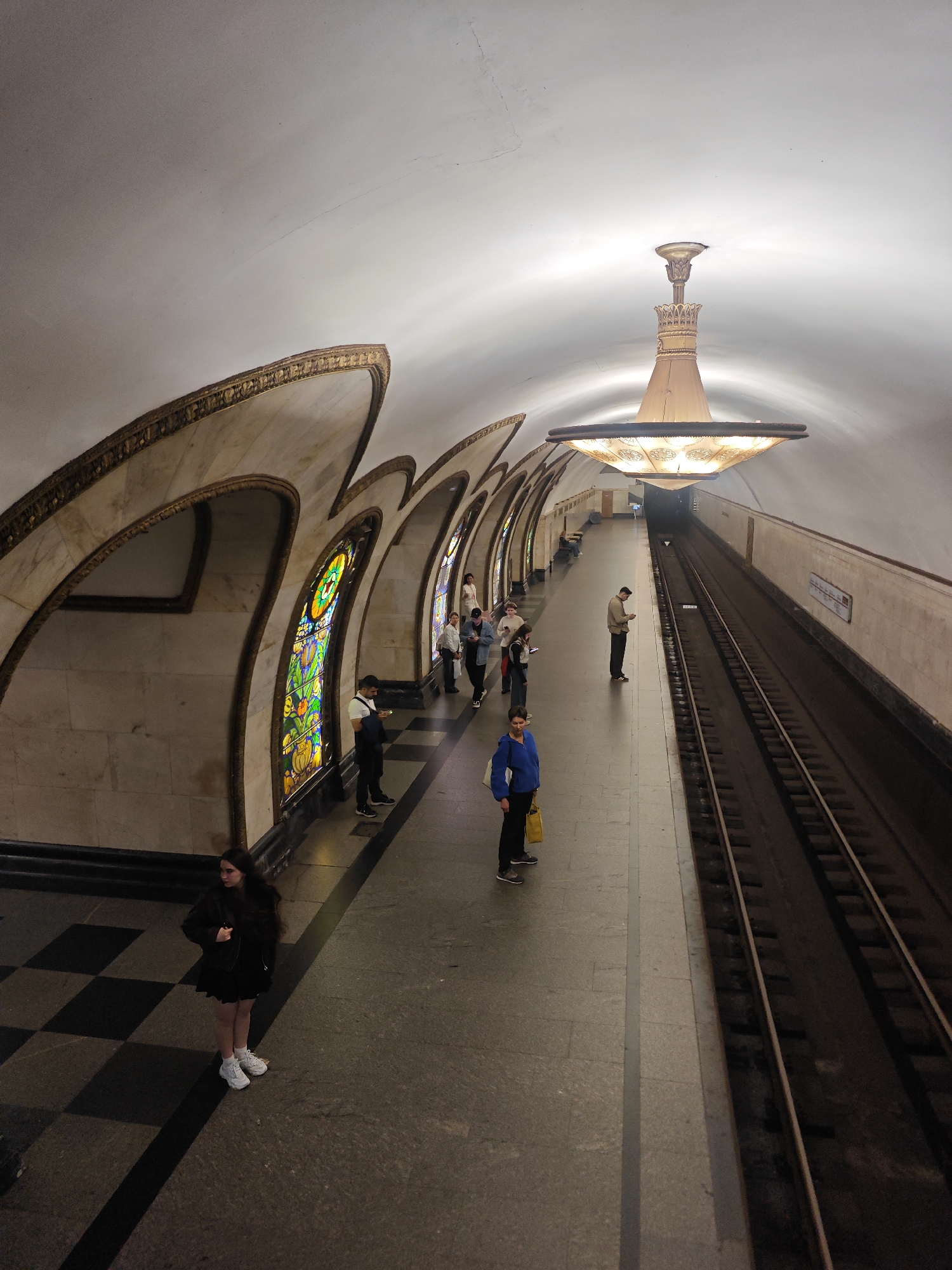 The image depicts an interior view of a Moscow Metro station, specifically the station Novoslobodskaya. The station's architecture is characterized by its Art Deco style, featuring a high, vaulted ceiling with a central, ornate chandelier. The walls are adorned with marble and decorative elements, including arched niches that house stained-glass windows.
On the platform, several individuals are visible, standing or walking. In the foreground on the left, a young woman with long dark hair is standing, wearing a black dress and white sneakers. Further down the platform, a person in a blue jacket and black pants is holding a yellow bag. Other passengers are seen scattered along the platform, some looking at their phones.
The train tracks are clearly visible to the right of the platform. The overall atmosphere is dimly lit, with the light emanating from the chandelier and other fixtures creating a sense of grandeur and depth. The floor of the platform features a distinctive checkered pattern. The station's unique design and the presence of passengers suggest an active transit environment.