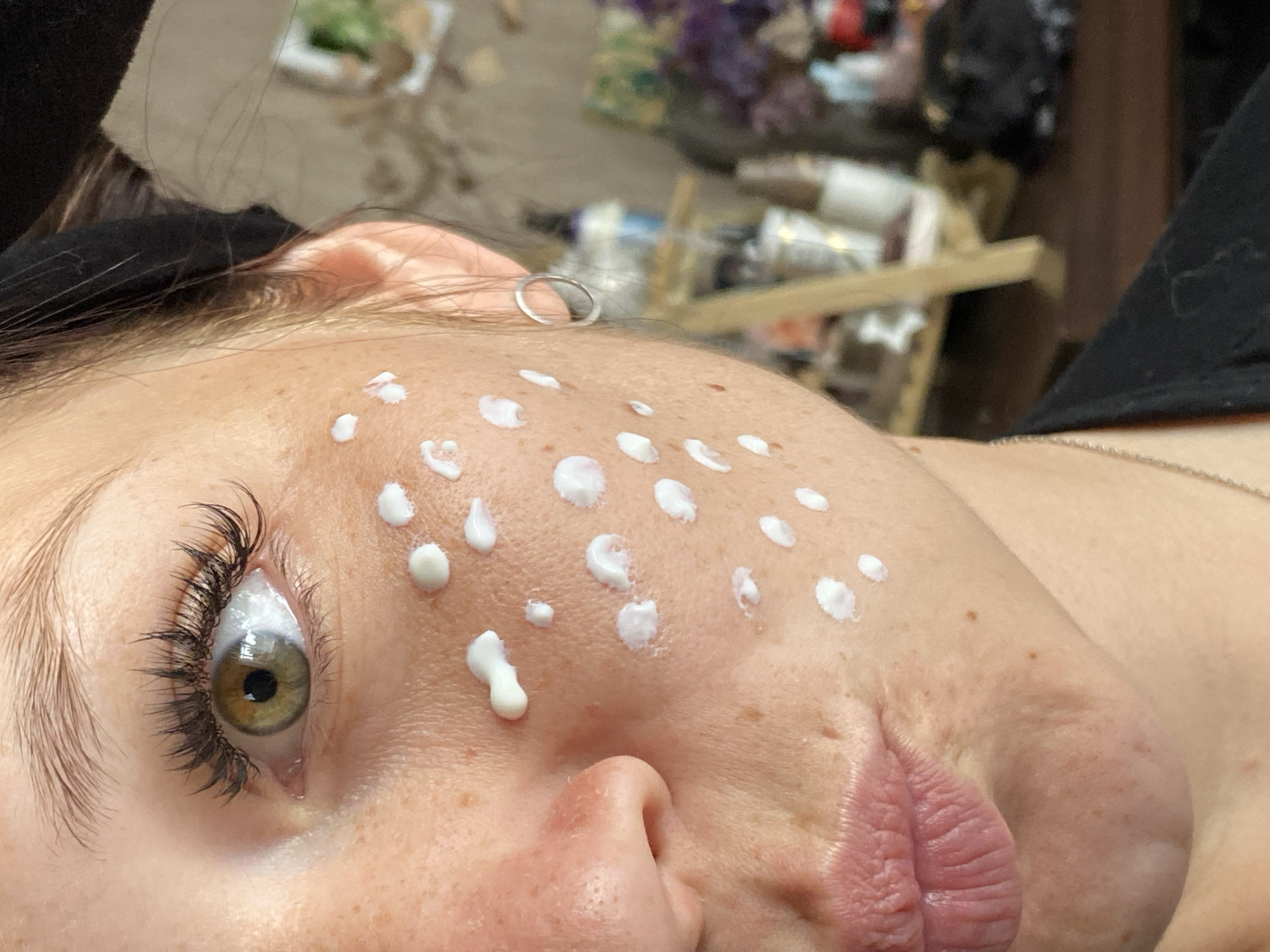 The image is a close-up, eye-level, outdoor shot of a young Caucasian woman's face, primarily focusing on the left side. She has fair skin with freckles, green eyes, and dark eyelashes. White dots of a cosmetic product are applied to her cheek and forehead. She is wearing a black headband that partially obscures her hair. Her mouth is slightly pursed, and she is wearing a thin silver hoop earring in her left ear. The background is blurred but appears to be an indoor setting with shelves containing bottles and other items, including what looks like a vase of purple flowers on the right. There is no visible indication of the city of Chaikovsky, Russia, or any specific urban environment.
