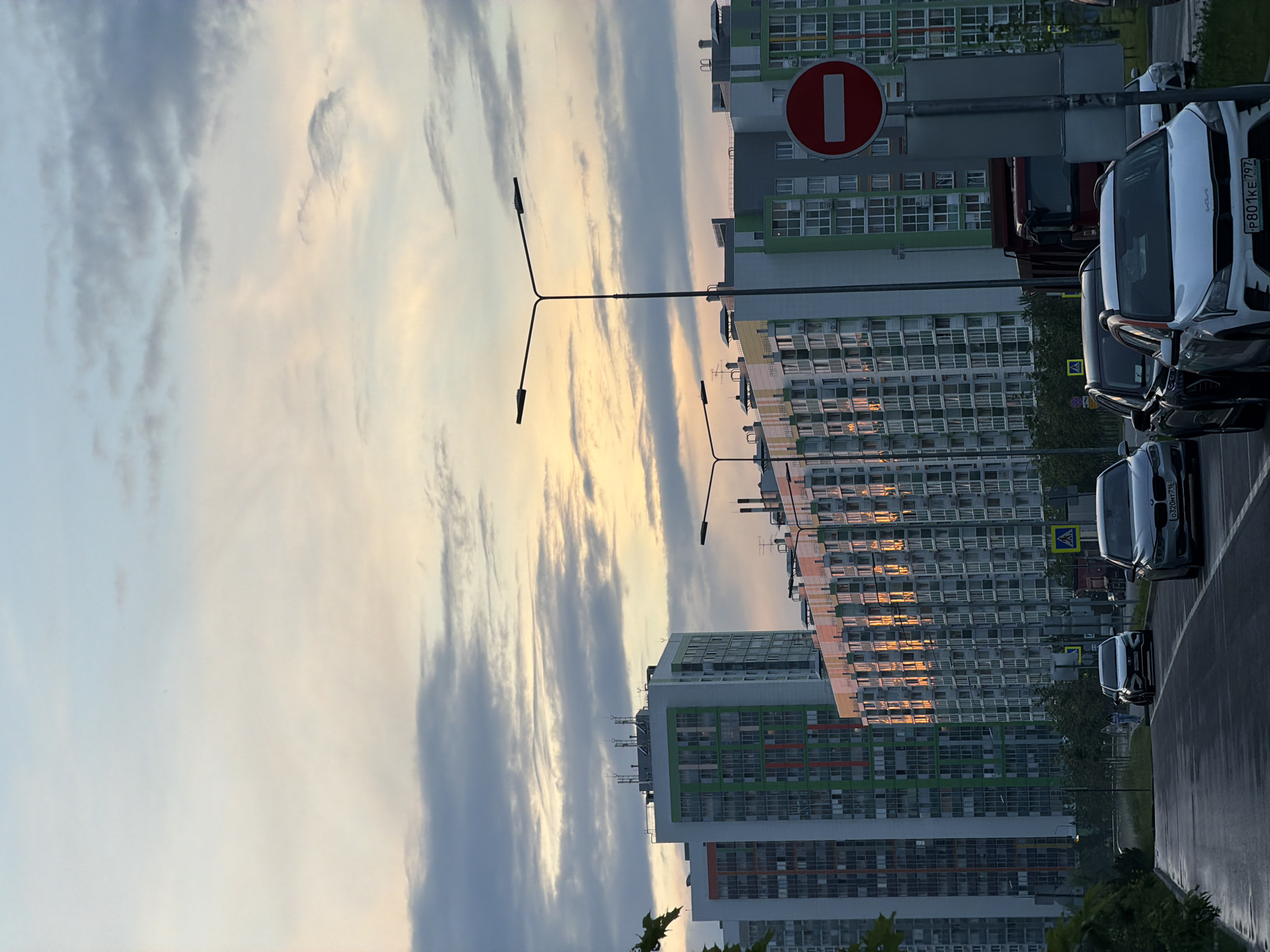 In a cityscape at dusk, a street is lined with tall, modern apartment buildings with colorful facades. The sky is filled with dramatic clouds, with a soft, warm glow of sunset visible through them. Several cars are parked on the wet pavement of the street, and a red truck is visible in the background. A "no entry" sign is mounted on a pole on the right side of the image. The scene is quiet and still, with a few figures walking in the distance.