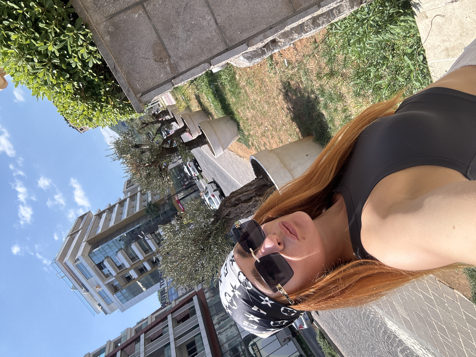 The image is a selfie taken outdoors, likely in an urban environment given the presence of modern buildings. The subject is a woman with long, reddish-blonde hair, wearing sunglasses and a black headscarf with a repeating pattern of LV logos and white floral designs. She is also wearing a black athletic top. The image is taken from a slightly high angle, looking down towards the subject.
In the background, there are contemporary buildings with glass facades and balconies. There are also trees with visible bark and foliage, and a patch of grass. Some of the buildings appear to be under construction or recently built. The sky is blue with scattered white clouds, suggesting a daytime scene. The lighting indicates it is sunny.
There is some visible text on the buildings and on signs, but it is not clearly legible in the provided image. One sign appears to say "Market". The overall impression is one of a sunny day in a modern city, possibly in Budva, Montenegro, as indicated by the location context. The subject's pose and attire suggest a casual or possibly athletic outing. There are no overt displays of emotion, but the selfie format and sunglasses lend an air of casual confidence. The scene is composed of a mix of natural elements like trees and grass, and man-made structures like buildings and paved walkways.