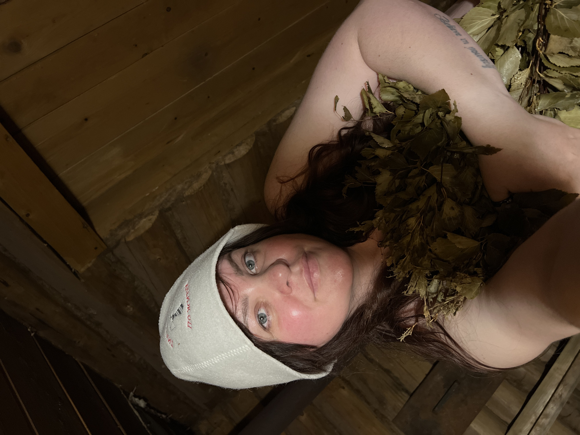 A woman lies among the leaves in what appears to be a sauna. She is fair-skinned with brown hair and blue eyes, wearing a white sauna hat with red lettering. There is a tattoo that says "Believe in yourself" on her arm. The background shows wooden planks, typical of sauna interiors. The image was taken in a sauna, and the presence of the sauna hat and leaves suggests the woman is engaging in a traditional sauna ritual. There is no indication this image was taken in the city of Protasy, Russia.