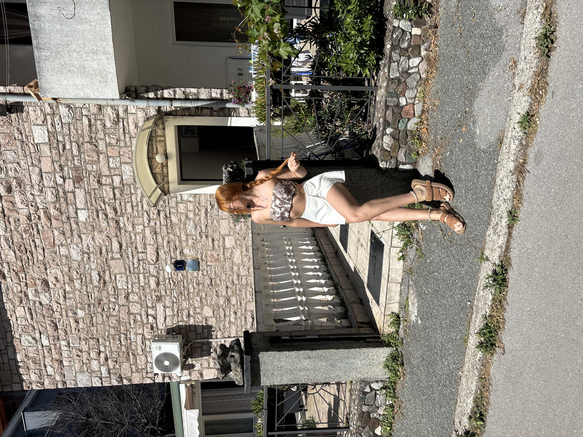 A young woman with long, braided red hair stands in direct sunlight, leaning against a low stone wall and a grey granite pillar at the entrance of a residential property in Budva, Montenegro. She wears a strapless, light-colored crop top with a textured or patterned design, a white wrap-style mini skirt, and brown fuzzy slide sandals with pearl-like ankle bracelets on both ankles. Her left hand lightly grasps her braid, and her gaze is directed towards the left side of the frame, partially obscured by her hair.
The property features a multi-story building with a light reddish-brown natural stone facade. Mounted on the stone wall is a white FOX brand air conditioning unit. Below it, a blue circular sign displays the number '1', and beneath that, a rectangular sign reads "APARTMAN". The main entrance, visible behind the woman, has a light-colored arched awning. To the right of the entrance, a grey downspout runs vertically along the building, and a small wooden birdhouse is affixed high on the wall, near a white stucco balcony.
A white balustrade fence and a black metal gate separate the property's tiled courtyard from the asphalt sidewalk. The granite pillar, topped with a grey rock formation, marks the edge of the gate. Lush green foliage, including bushes and a vine, grows within the property behind the black fence. The foreground shows a grey asphalt road with a curb and patches of weeds growing along the edges of the pavement and curb. The scene is brightly illuminated, indicating a clear, sunny day.
