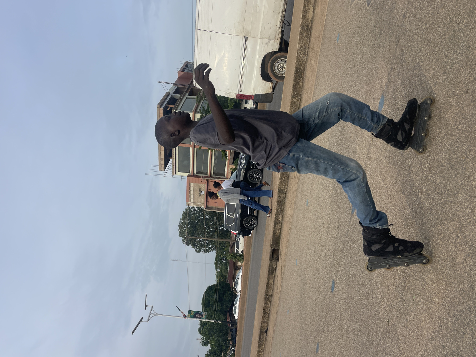 In Jos, Nigeria, a young boy with dark skin, wearing a dark grey t-shirt and blue jeans, is rollerblading on a paved road. He is captured in profile, facing right, with his right arm extended forward and his left arm bent at the elbow. He is wearing black rollerblades. In the background, a white van is parked on the right side of the road, and several cars are visible further down the street. Two individuals, a woman and a man, are walking on the sidewalk on the left side of the road. Buildings and trees line the background, and streetlights are also present. The sky is overcast.