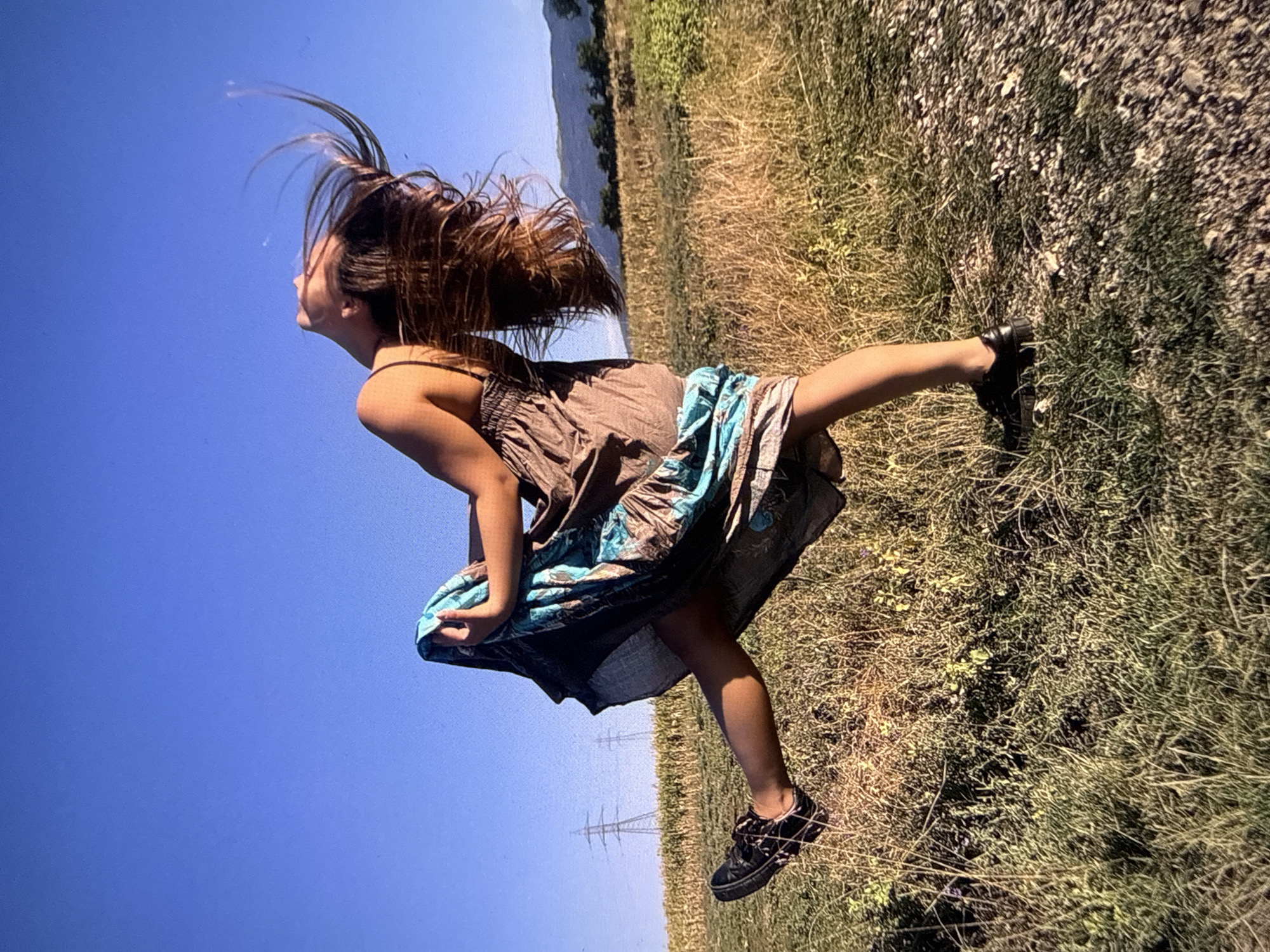 A young woman in a brown and blue dress is captured mid-movement outdoors. She is positioned on a grassy field under a clear blue sky. Her brown hair is thrown back. She is wearing black shoes. In the background, there are power lines and a distant view of a river or lake. The image is angled, creating a tilted perspective. The location is suspected to be in Sofia, Bulgaria.