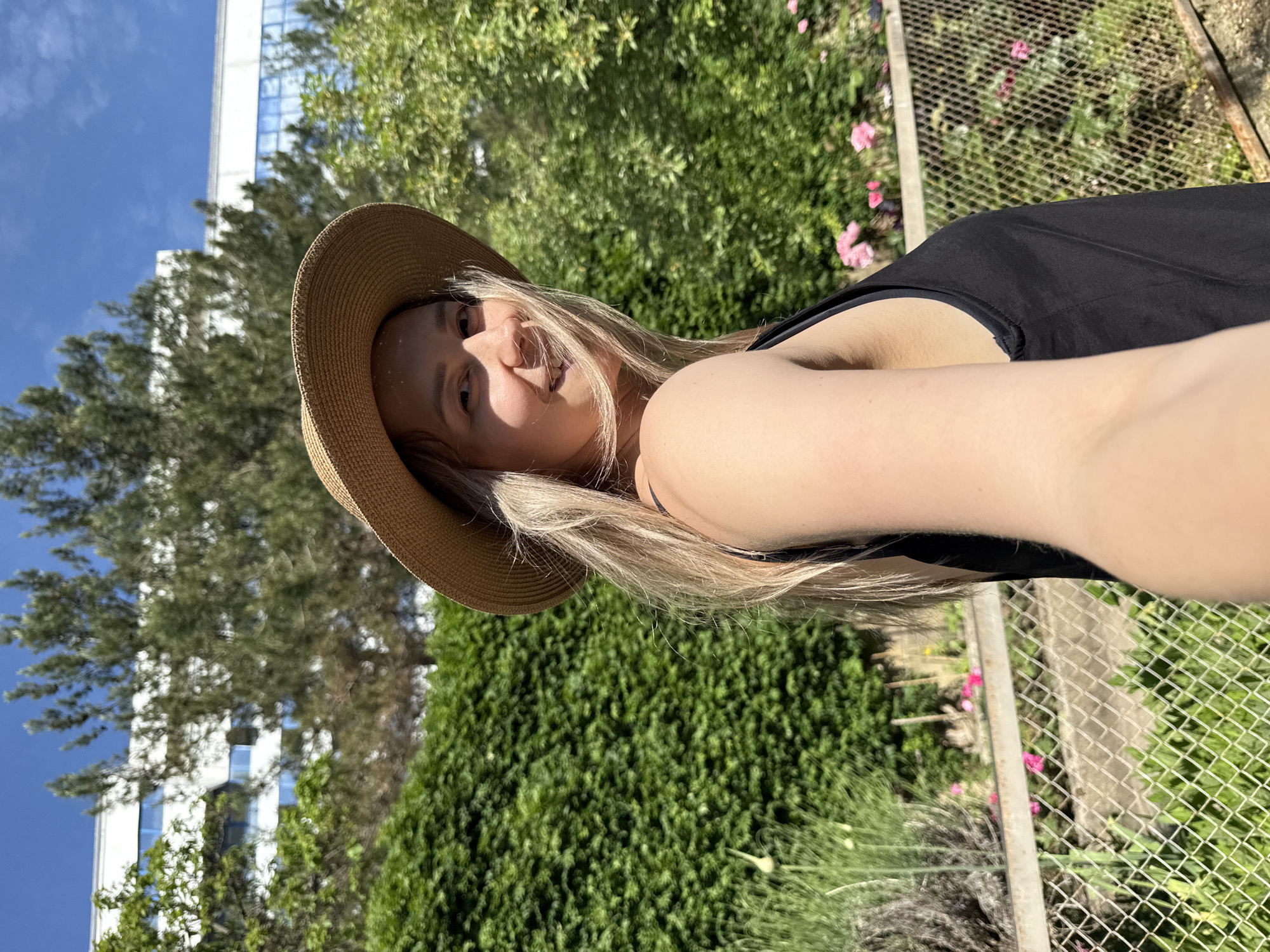 In the city of Nebug, Russia, a young woman with long blonde hair and a straw hat is captured in a selfie. She is wearing a black tank top and appears to be outdoors, with a lush green hedge in the background. A modern building with white and blue accents is visible behind the foliage, and a chain-link fence runs across the foreground. The scene is illuminated by bright sunlight, suggesting it is daytime.