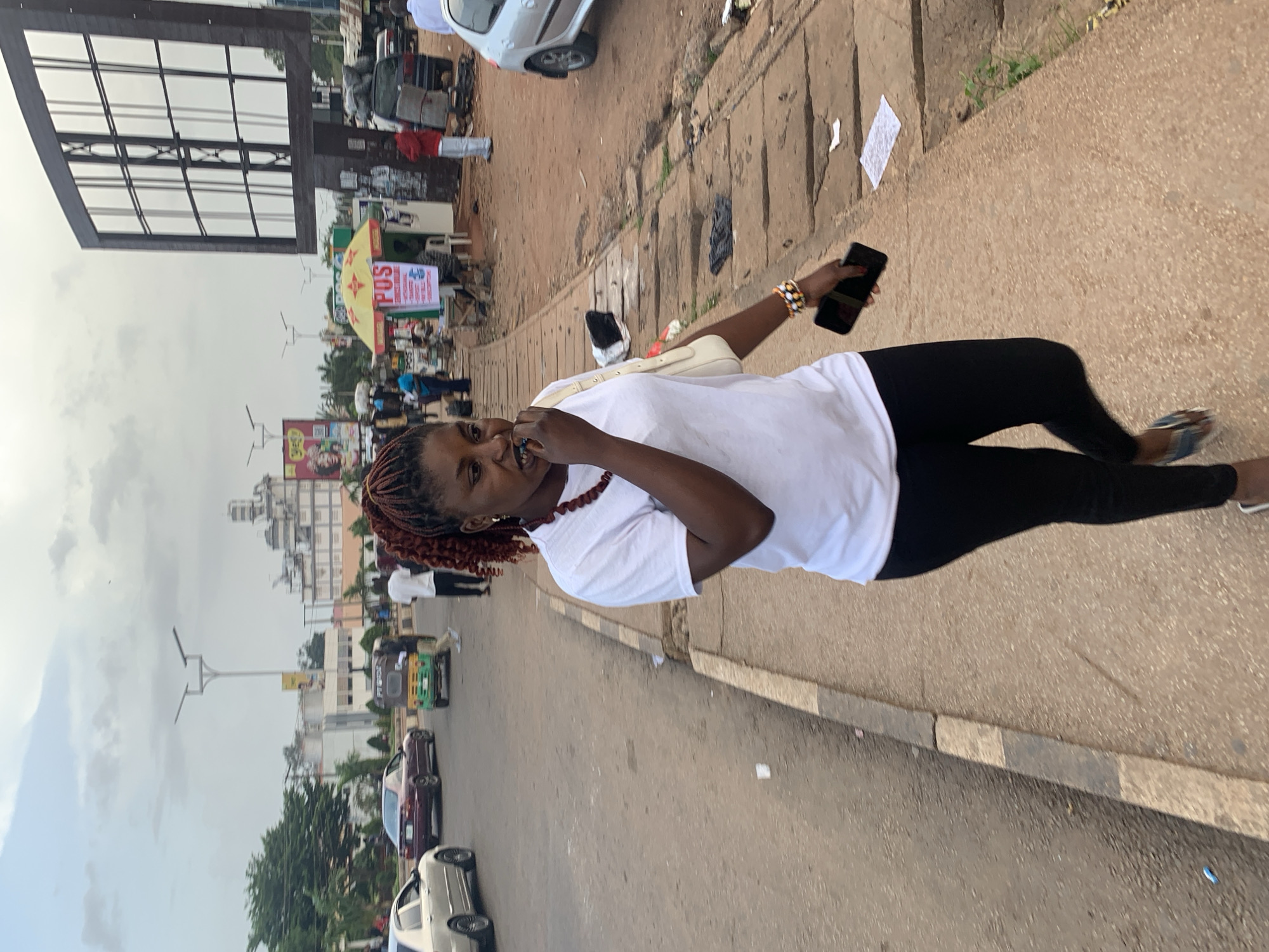 In Jos, Nigeria, a young woman with braided reddish-brown hair walks on a sidewalk next to a busy street. She is wearing a white t-shirt, black leggings, and patterned sandals. A white purse is slung over her shoulder, and she holds a black smartphone in her right hand, bringing it to her mouth. She is wearing a beaded necklace and bracelets. The street is filled with vehicles, including cars and a three-wheeled auto-rickshaw. Several businesses, including a POS service stall with a yellow umbrella, are visible along the sidewalk. A large billboard stands prominently in the background. The sky is overcast, suggesting cloudy weather.