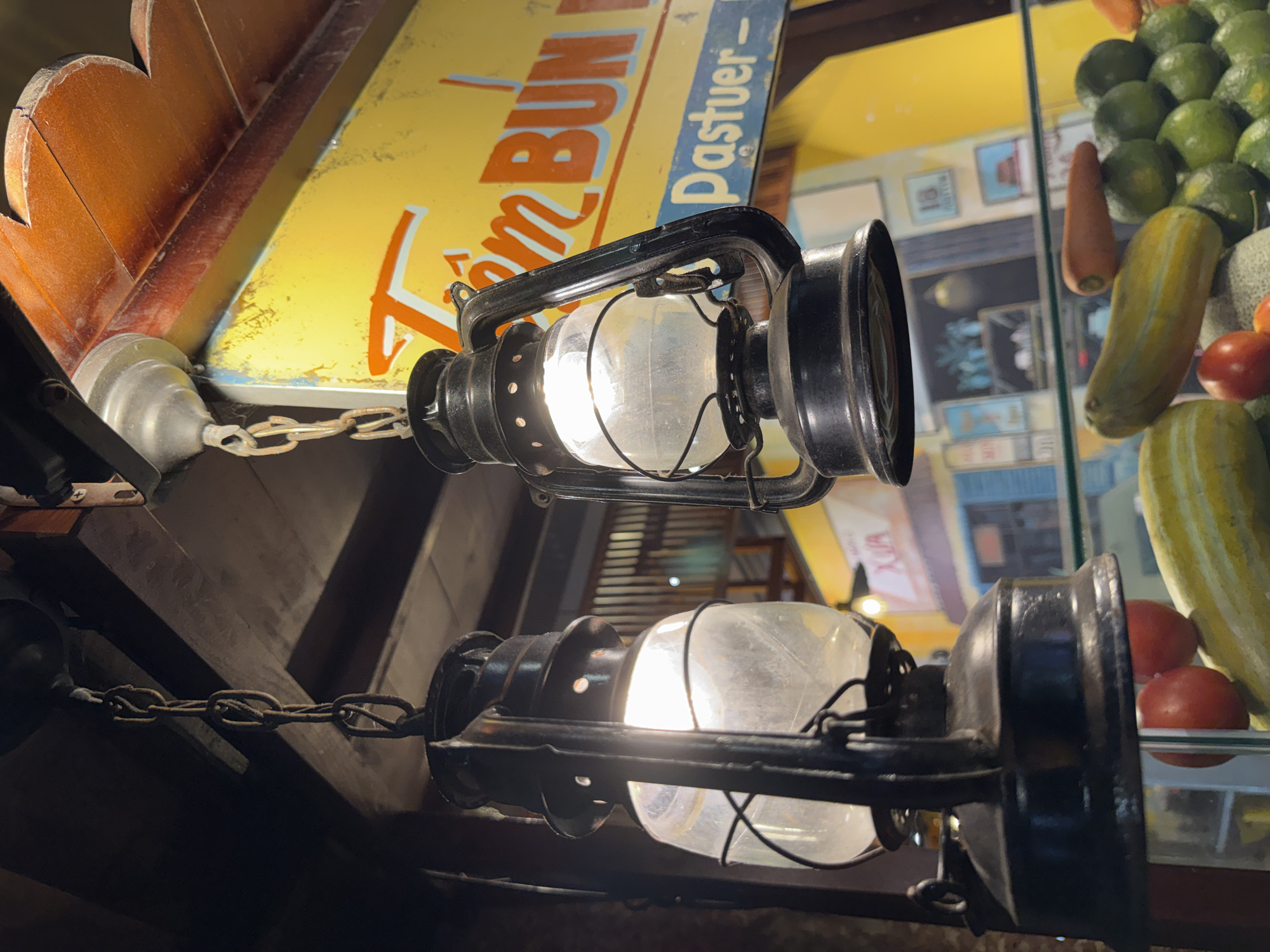 This media file captures a close-up view of two vintage oil lanterns hanging in what appears to be a market or food stall in Nha Trang Ward, Vietnam. The lanterns, made of dark metal with glass globes, are illuminated, casting a warm glow. They are suspended by rusty metal chains, suggesting an aged and rustic aesthetic.
In the background, a yellow sign with red and blue lettering is visible, with partial text reading "om BUN" and "Pastuer -". This likely indicates the name of a dish or a product being sold, possibly related to Vietnamese cuisine. Further in the background, through a glass display, a variety of fresh produce can be seen, including green limes, tomatoes, carrots, and what appear to be cucumbers or other gourds. The overall setting suggests a lively, perhaps evening or indoor, market environment. The lighting and the vintage lanterns evoke a sense of nostalgia and local culture. There are no people directly visible in the frame, but the setting implies a place of commerce and activity.