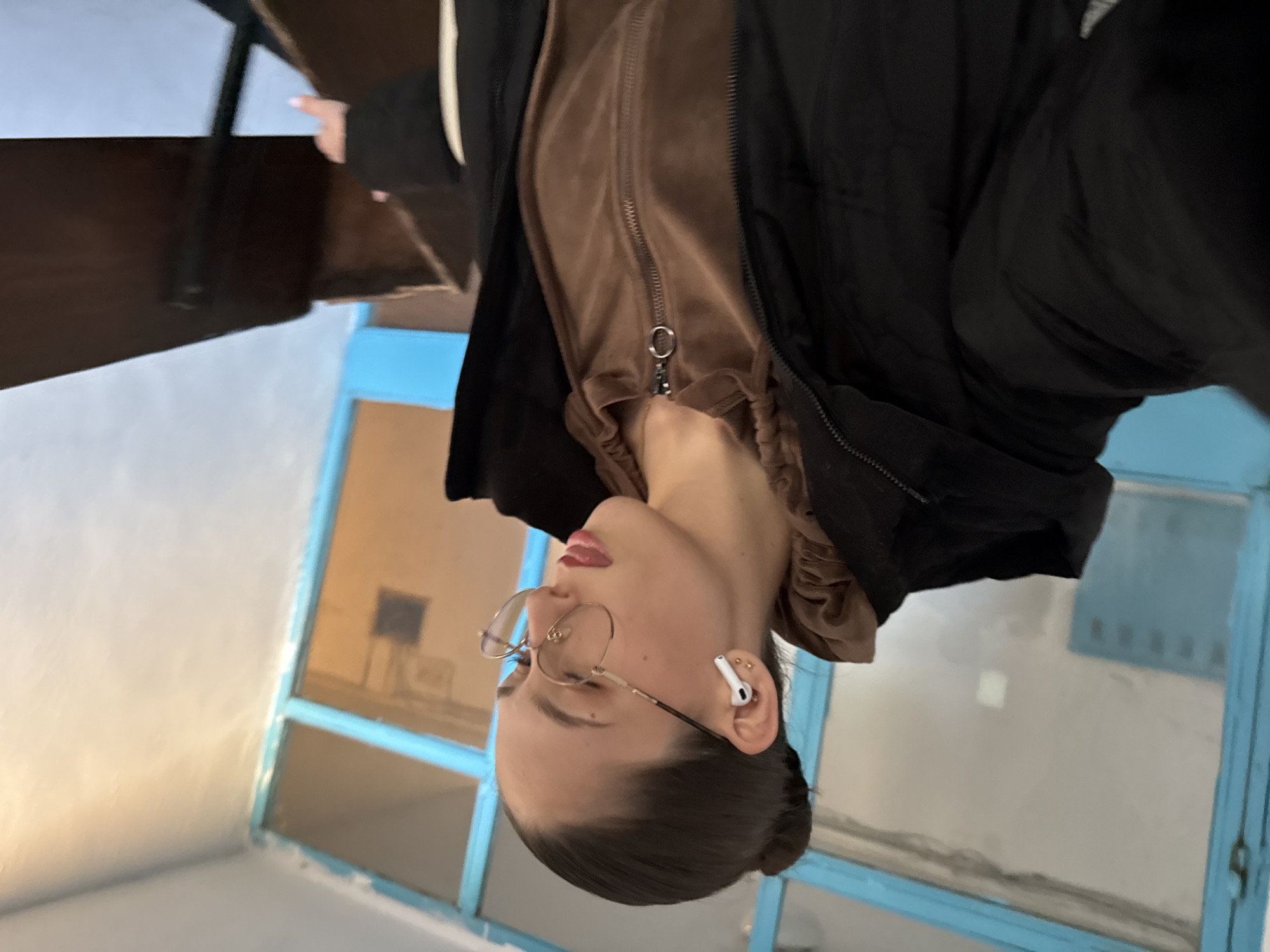 A woman with light skin, dark hair pulled back in a bun, and glasses is visible from the chest up, wearing a brown velvet zip-up jacket under a black puffer jacket. She has an AirPod in her right ear.
The location appears to be indoors, next to a window with a blue frame. Outside the window, there's a building with multiple windows. Based on the architectural style and color palettes, the image potentially captures a scene in Sofia, Bulgaria. Additional visual data would be necessary for verification.