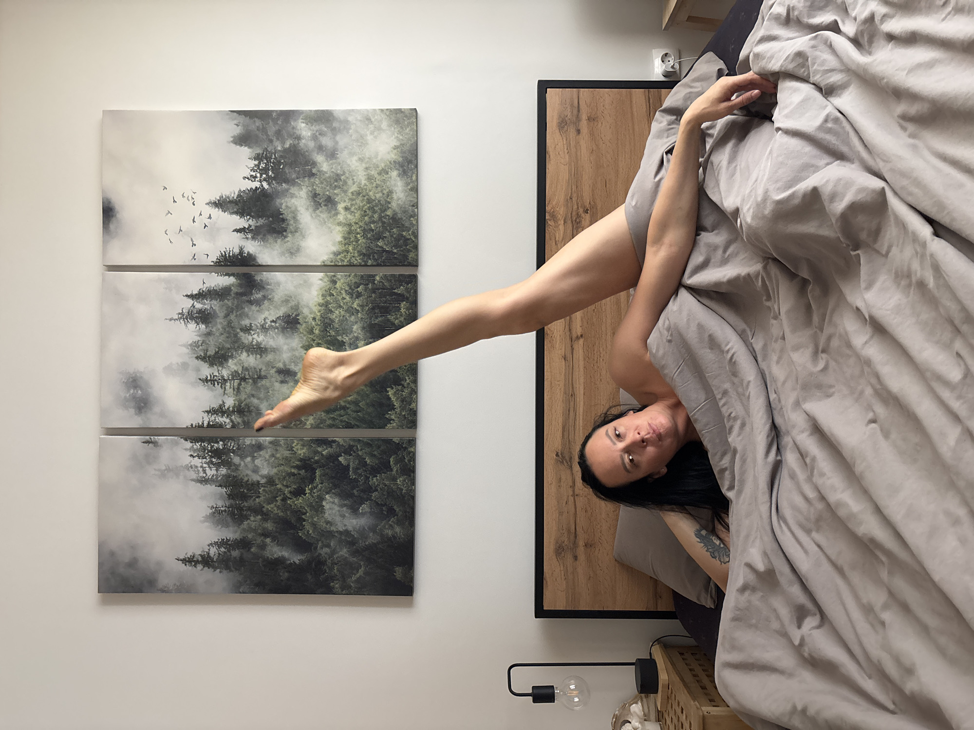 A woman is lying in bed, with one leg extended vertically upwards towards a three-panel artwork depicting a forest scene with mist and birds. She is partially covered by a grey duvet. The bed has a wooden headboard with a black frame. The artwork hangs on a white wall. To the side of the bed, there is a small bedside table with a black lamp. The image was taken in Chaikovsky, Russia.