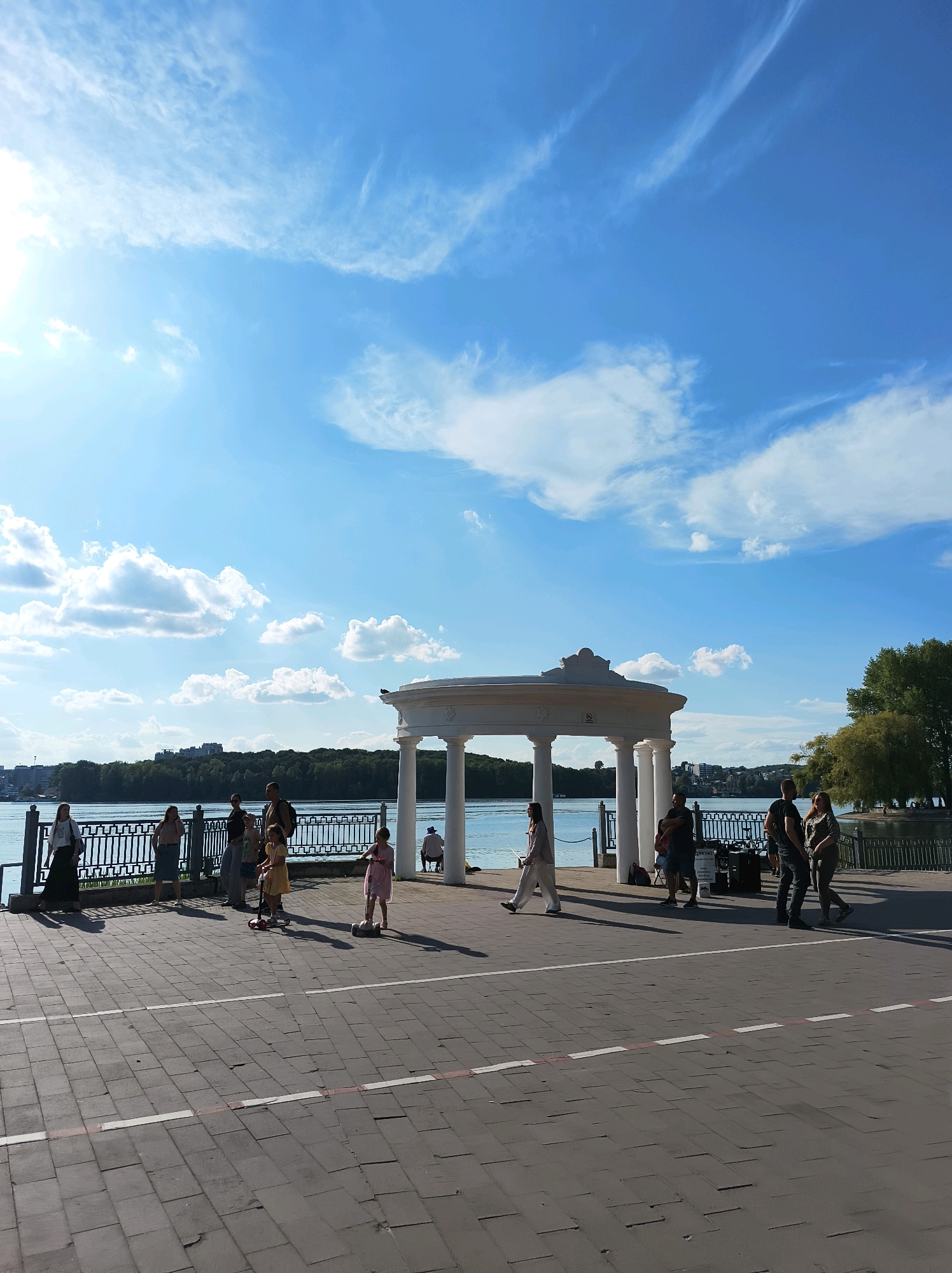 The image depicts a sunny day along the Ternopil Lake promenade in Ternopil, Ukraine. A white, open-air gazebo with a domed roof and classical columns stands prominently on the right side of the frame. In the foreground, a paved walkway is visible, marked with white lines. Several people are present on the promenade. On the left, a woman in a light-colored outfit walks away from the camera, and further along the railing, a group of people stands conversing. Two young girls are on scooters in the middle of the promenade, one on a pink scooter and the other on a white one. On the right side, near the gazebo, a group of adults are standing and talking. The lake is visible behind the promenade, with a tree-lined shore and buildings in the distance. The sky is bright blue with scattered white clouds.