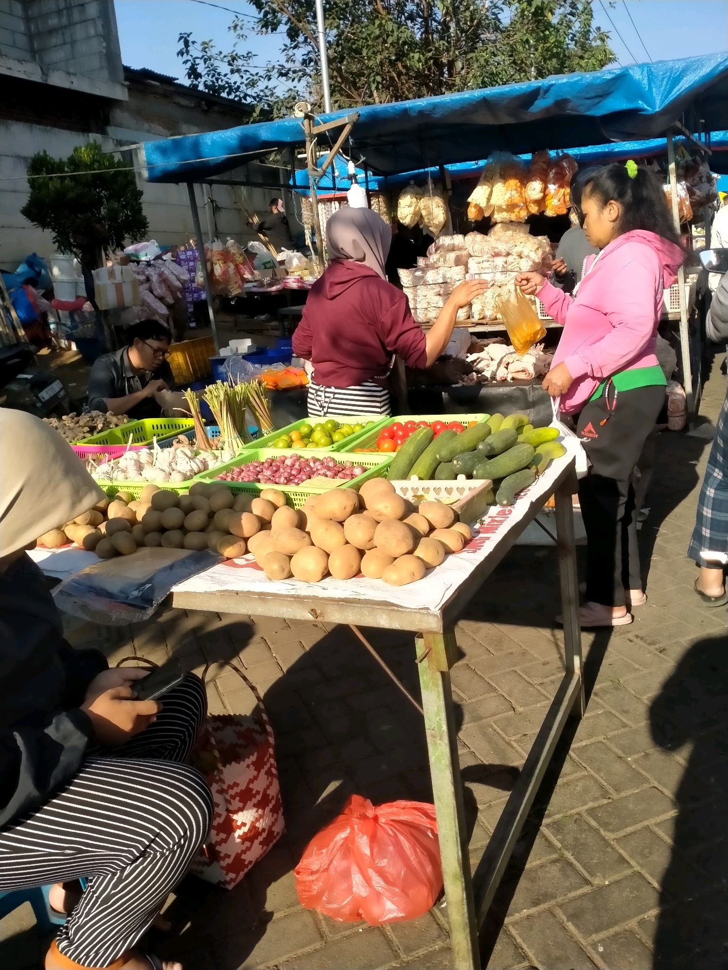 An outdoor market stall is captured in Batu, Indonesia, under a blue tarp awning. In the foreground, a person wearing a headscarf and a maroon hooded sweatshirt is reaching towards another person. The second person, wearing a bright pink hooded sweatshirt and black pants with a green waistband, is holding a plastic bag with yellow contents. They appear to be engaged in a transaction.
The stall displays a variety of fresh produce, including potatoes, garlic, onions, limes, tomatoes, and cucumbers, arranged in green plastic crates on a metal table. Behind the produce, several bags of snacks are hanging. To the left, a man wearing glasses and a dark shirt is seated, looking down at a bag in his hands. The ground is paved with interlocking bricks, and sunlight casts shadows. In the background, a building and trees are visible. A red plastic bag and a patterned woven bag are on the ground near the table.