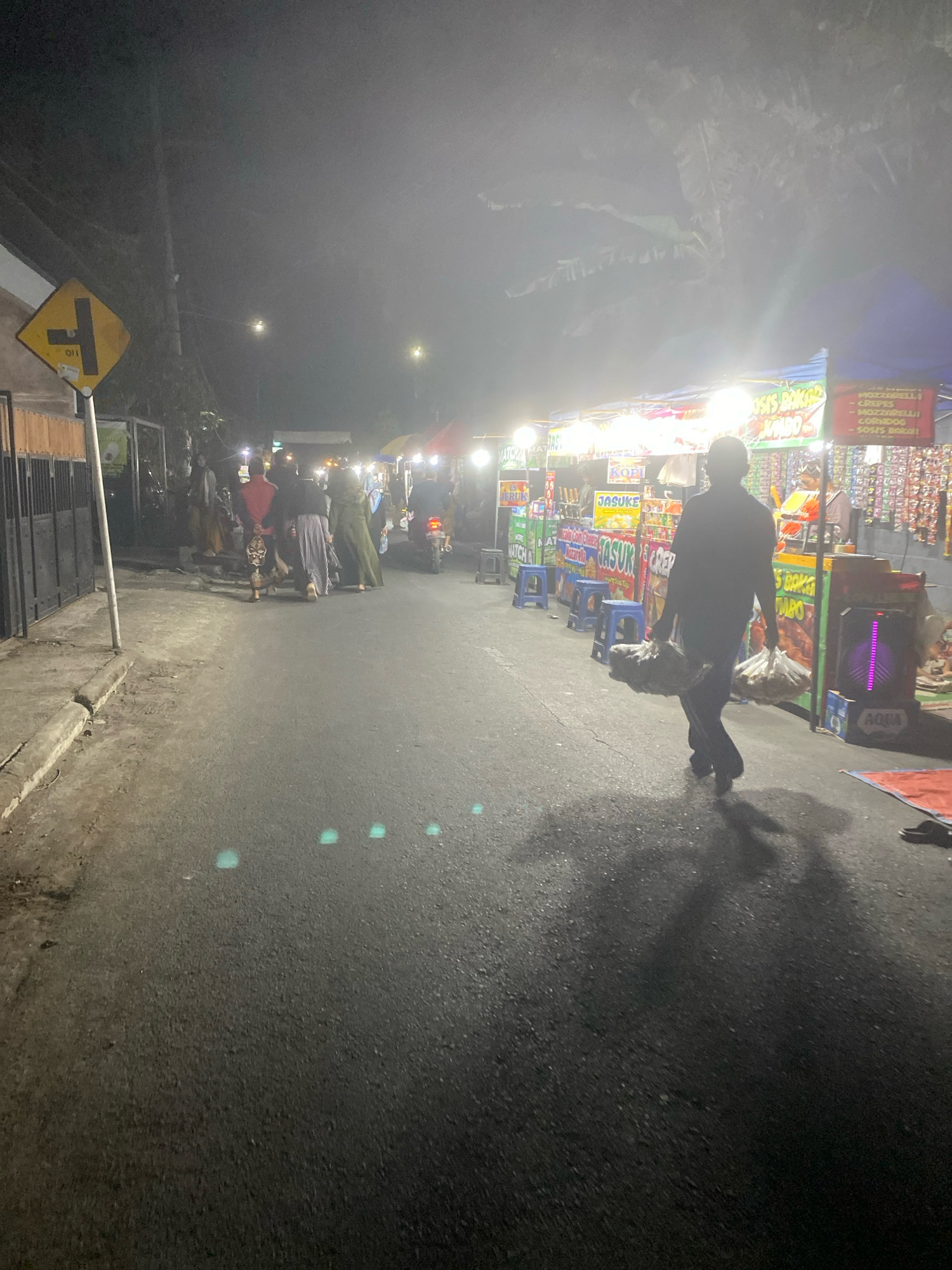 An exterior, nighttime, street-level shot depicts a hazy, dimly lit scene of a night market or food stall area in Kediri City, Indonesia. The street is paved, with a slight incline. On the left, a metal gate borders a property. Further left, a yellow diamond-shaped traffic sign with a black symbol indicates a T-junction. A sidewalk or raised curb runs along the left side of the street.
Several people are visible, mostly indistinct due to the low light and haze. On the left, a group of individuals, some appearing to be women in traditional clothing, are standing or walking. In the center right, a man is walking away from the camera, carrying two plastic bags filled with items. He is silhouetted against the lights of the stalls. Behind him, other figures are seen near the stalls.
The right side of the street is lined with various food stalls, indicated by colorful awnings and bright lights. Signs with Indonesian text are visible above the stalls, advertising food items. Several blue plastic stools are placed in front of one stall. An electronic device with purple lights, possibly a speaker or cooler, is situated near one of the stalls. The overall atmosphere suggests a bustling, yet somewhat murky, urban night market.