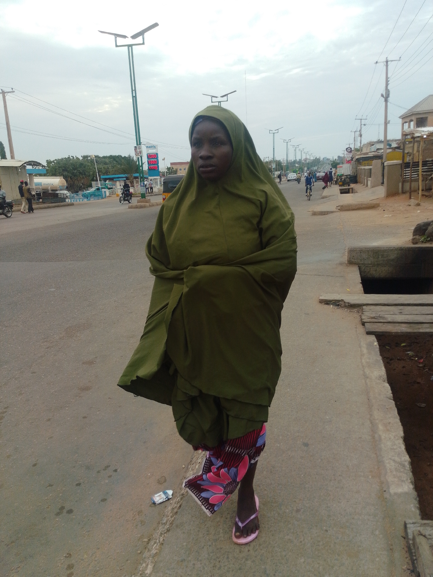 The image depicts a street scene in Gombe, Nigeria. In the foreground, a woman wearing a green hijab and a patterned skirt walks on a paved sidewalk. She is facing the camera and is partially cropped out of the frame. Her left foot, adorned with a pink flip-flop, is visible. To her right, a concrete drainage channel runs along the edge of the sidewalk, partially covered by wooden planks.
The street is paved with asphalt and appears to be a busy area with traffic. Vehicles, including cars and motorcycles, are visible in the background. A gas station with a prominent "Matrix" sign and a price display showing fuel costs is located on the left side of the street. Streetlights with solar panels are installed along the road. Buildings of various sizes and styles line the street, some appearing to be shops or residences. Power lines are strung overhead. The sky is overcast with clouds. A small toy car is lying on the sidewalk near the woman's feet.