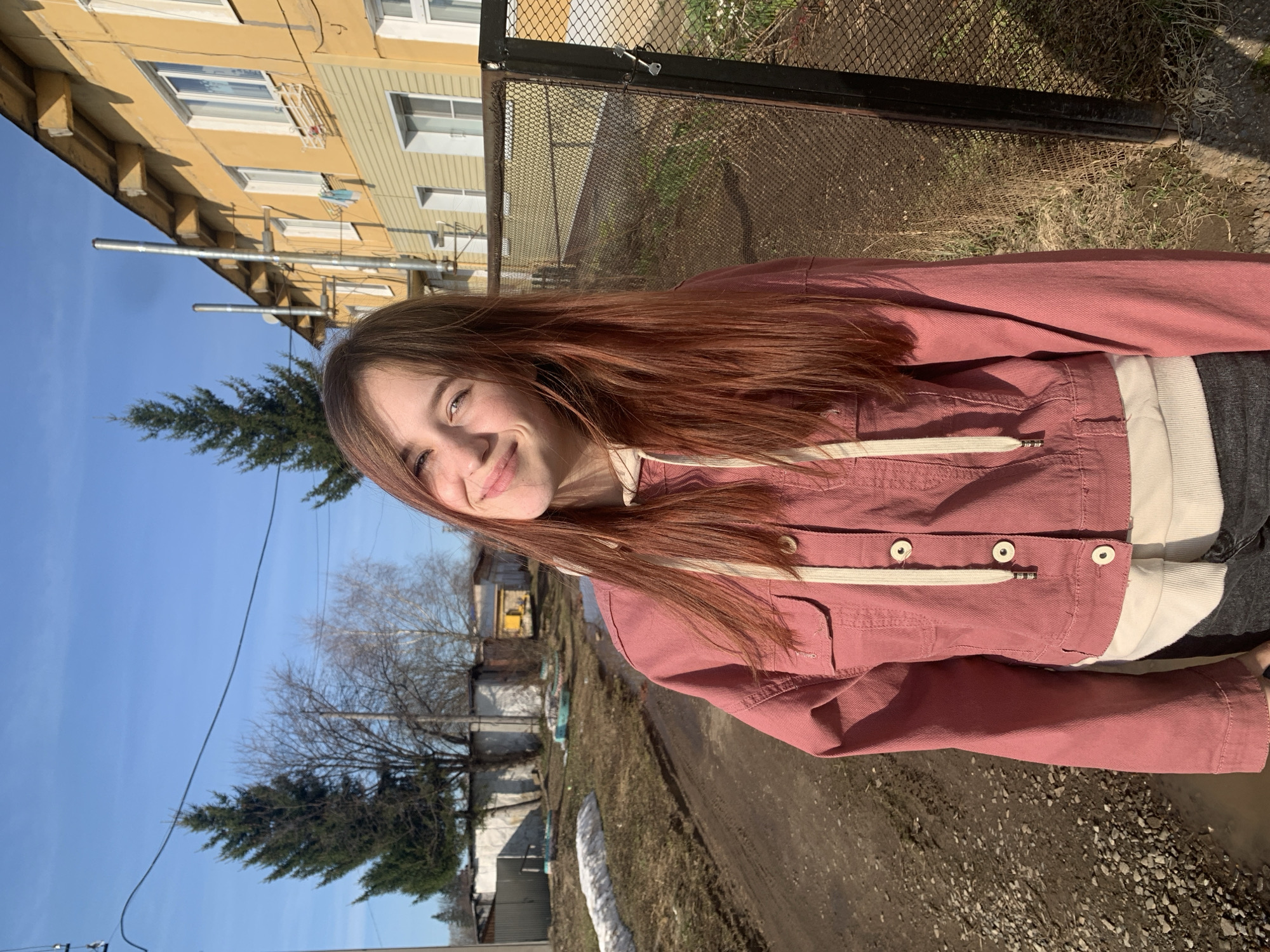 A young woman stands on a dirt road in what appears to be a suburban or rural area, smiling towards the camera. She is wearing a pink jacket over a white hoodie, and dark-colored pants. Behind her, there are houses and trees, along with a fence. The sky is blue. The photo seems to be taken outdoors during daytime. Based on the information provided, the location is Фоки, Russia.