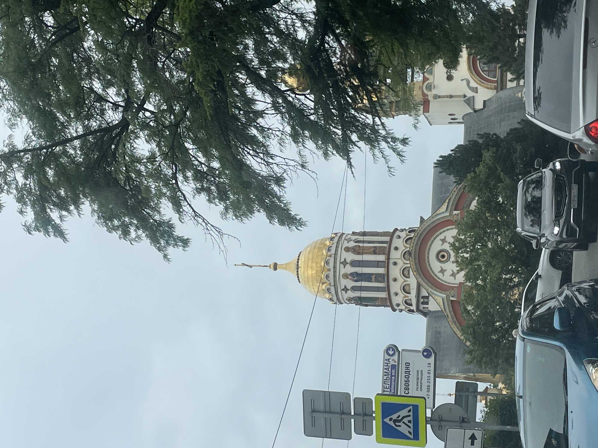 A golden-domed cathedral with white and red architectural elements stands behind a street scene with traffic and pedestrian signs. The cathedral features arched windows with figures depicted inside and a cross at its apex. In the foreground, several cars are visible, including a dark-colored SUV with a license plate and a silver sedan. A blue car is partially visible on the left. Pedestrian crossing and directional signs are mounted on poles. Dense green trees partially obscure the view of the cathedral and sky. The sky is overcast. The scene is likely on a cloudy day. This location is the Cathedral of the Archangel Michael in Sochi, Russia.