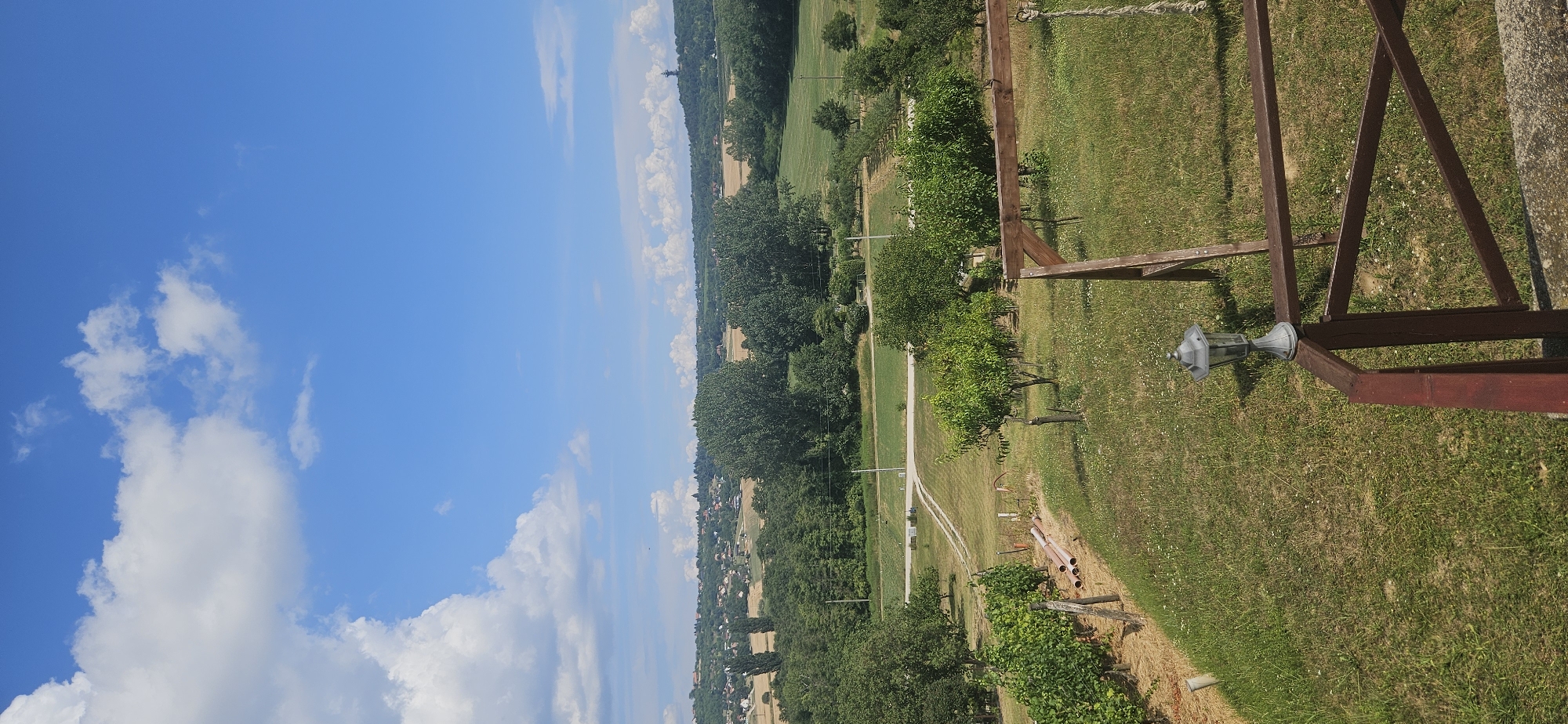 A high-angle, full outdoor shot captures a scenic view of the Hungarian countryside under a bright blue sky with fluffy white clouds. In the foreground, a grassy area with scattered small white flowers is bordered by a wooden railing and a lamppost. To the left, a vineyard with staked rows of green vines is visible, along with some orange pipes.
Beyond the foreground, the landscape transitions to a park-like setting with several young trees and a dirt path winding through the grass. Further in the distance, a dense line of green trees creates a natural border. Beyond this tree line, a village or town is nestled on rolling hills, with numerous houses featuring red-tiled roofs. Agricultural fields, appearing golden and green, are interspersed with the vegetation and the buildings. On the far right horizon, a distant tower or monument is visible on a hilltop. The overall impression is one of a serene rural environment.