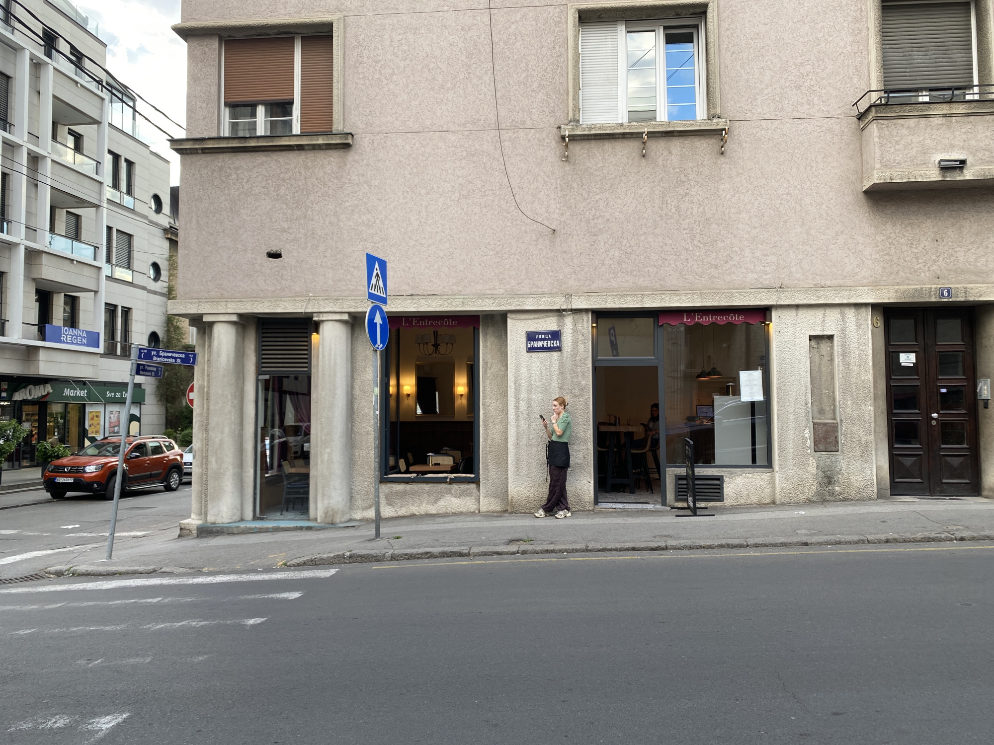 The image shows a street scene in Belgrade, Serbia, specifically the intersection of Branicevska Street and another street. On the left is a multi-story building with a storefront labeled "AROMA Market." An orange Dacia Duster car is parked on the street in front of the market. A street sign indicates that the cross street is Branicevska Street. The main focus is on the corner building, which houses two establishments, both named "L'Entrecôte." A woman stands on the sidewalk in front of one of the L'Entrecôte storefronts, using her mobile phone. The building's facade is beige, with architectural details such as pillars and window trim. There are two windows on the upper floors, one with a closed brown shutter and the other with an open white shutter. A black number 6 is visible next to the building's front door. The street is paved and shows signs of wear.