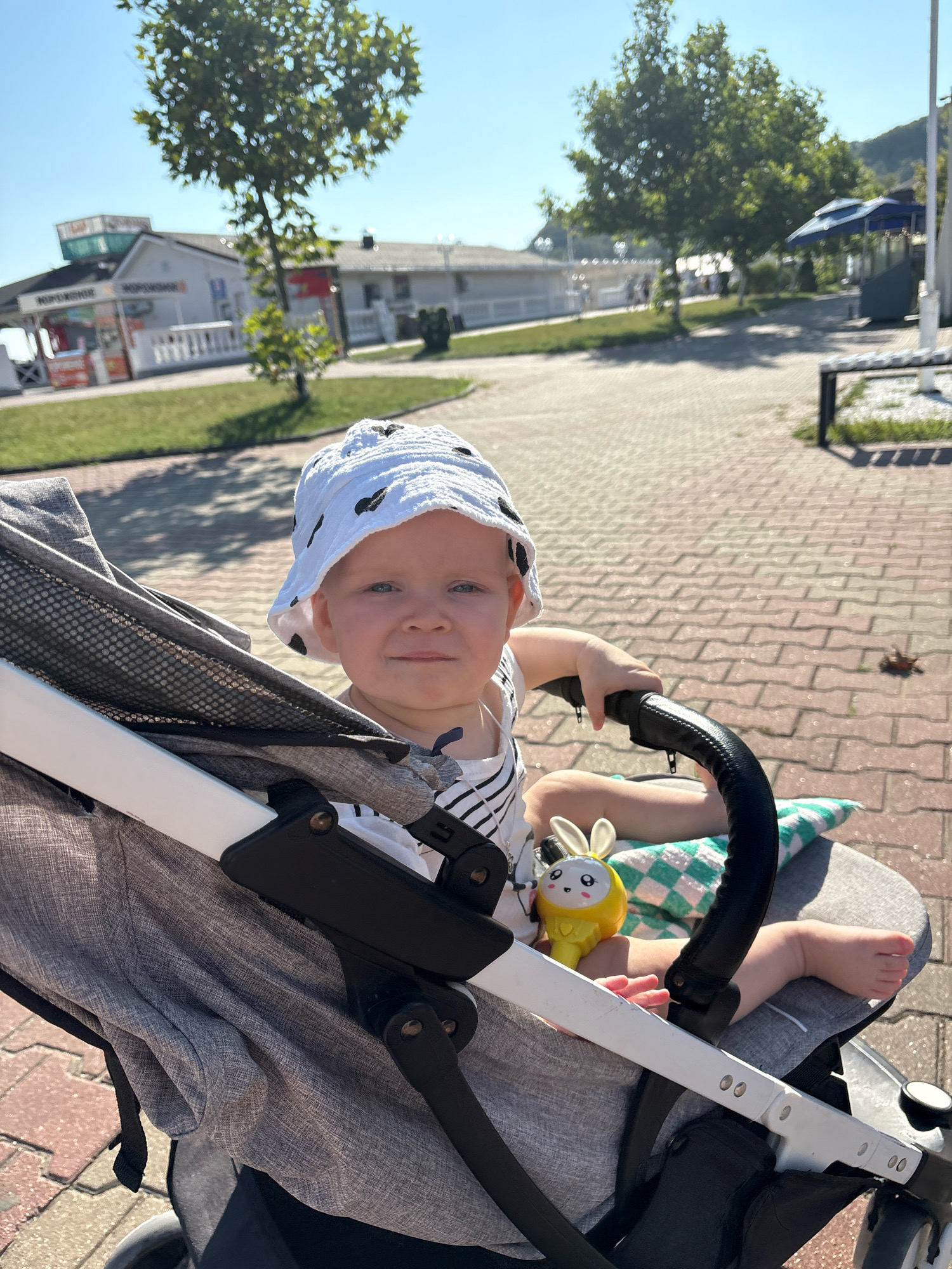 A close-up shot shows a young child, wearing a white bucket hat with black polka dots and a striped shirt, sitting in a stroller. The child is looking directly at the camera with a slight smile. In the stroller, next to the child, is a yellow bunny-shaped toy. A green and white checkered pillow is visible behind the toy. The stroller's frame is white and gray, with black handles.
In the background, a paved walkway with a brick pattern is visible, bathed in sunlight, suggesting it is daytime. Beyond the walkway, there is a grassy area with a few trees. Further back, several white buildings with red and green accents are situated, some appearing to be shops or kiosks. The text "ЖОРЖИЯ" is visible on one of the buildings. The overall scene depicts a sunny outdoor setting, likely a resort area or promenade in Arkhipo-Osipovka, Russia. The child appears content and comfortable in the stroller.
