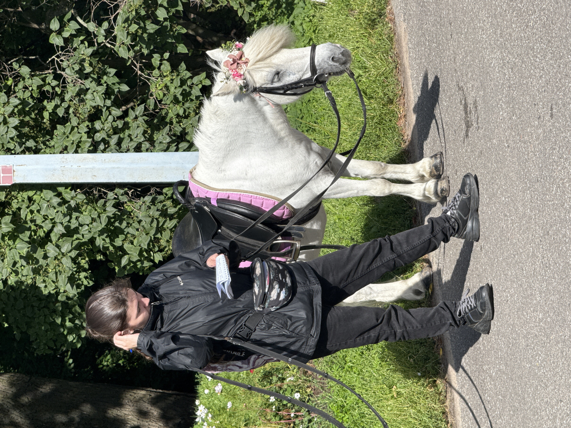 A young woman wearing a black jacket, black pants, and black sneakers stands next to a white pony. The pony is wearing a saddle pad with pink trim and a floral wreath on its head. The woman is holding a small notepad and appears to be writing or checking something in it. She has a black fanny pack around her waist. They are on a paved path next to a grassy area with some white flowers. Behind them is lush green foliage and a light blue pole with a red top, likely part of a park or street fixture. The scene appears to be outdoors in a park or recreational area, possibly in Saint Petersburg, Russia, given the context provided for the location.