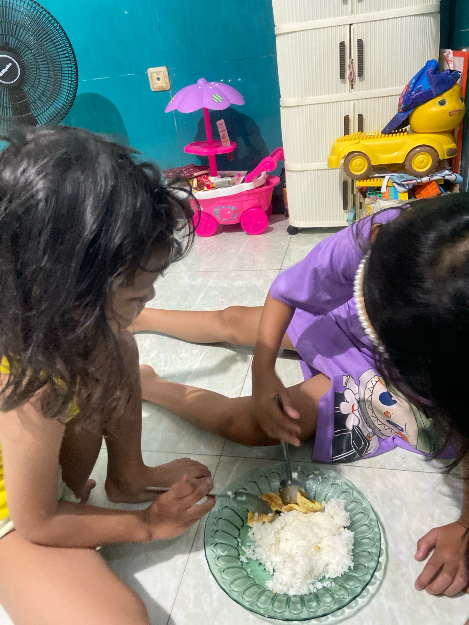 In a room in Gelam, Indonesia, two young children are sitting on a tiled floor, engaged in eating a meal. One child, wearing a yellow top, is on the left, facing away from the camera and towards the other child. Their legs are bent and tucked beneath them. The second child, on the right, is wearing a purple t-shirt with a cartoon character graphic and a pearl necklace. This child is also sitting with their legs bent, partially facing the camera. Both children are focused on a green glass plate filled with white rice and some pieces of scrambled egg, which they are eating with spoons.
The environment appears to be a child's room or play area, with toys visible in the background. To the left, a black oscillating fan is mounted on the wall, and a pink toy umbrella with a stand is also present. A tall, light-colored plastic storage cabinet stands against the back wall, with a yellow toy car and a blue cap placed on top of it. The wall behind the children is painted a bright teal color, and the floor is made of light-colored tiles. The lighting suggests an indoor setting, possibly with natural light supplemented by artificial sources. The overall scene depicts a casual mealtime moment for the children.