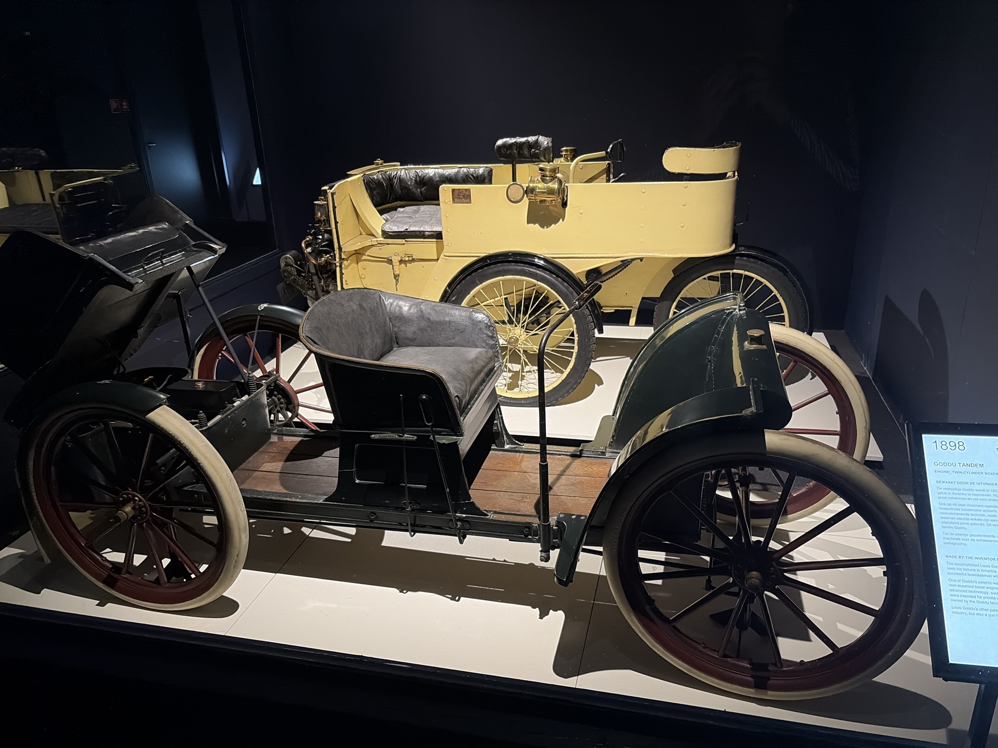 Two antique cars are on display in what appears to be a dimly lit museum or exhibition hall. The foreground vehicle is a dark green or black tandem-style car with a single grey leather bucket seat and wooden floorboards. It features large spoked wheels with dark red rims and light-colored tires. To its left, partially obscured, is another similar car. In the background, a light yellow or cream-colored car is visible, also featuring spoked wheels with yellow rims and black tires, and a black upholstered bench seat. Both vehicles are early automobiles, characterized by their open designs and prominent spoked wheels.
The setting is characterized by dark walls and a light-colored platform on which the cars rest, suggesting a curated display environment. Spotlights illuminate the vehicles, casting shadows on the platform and enhancing their historical presence.
To the right of the foreground car, a display sign provides information about the vehicles. Visible text on the sign identifies one car as:
- **1898**
- **GODDU TANDEM**
- **ENGINE: TWIN-CYLINDER BOXER**
The sign, partially in Dutch ("GEMAAKT DOOR DE UITVINDER") and English ("MADE BY THE INVENTOR"), details that the inventor, Louis Goddu, born in 1833, sought his fortune in America as a businessman. It mentions the "rear-mounted boxer engine" and "advanced technology" of the car, which was intended for private use and owned by the Goddu family. It also notes Goddu's other patents in the shoe industry and a gun.
