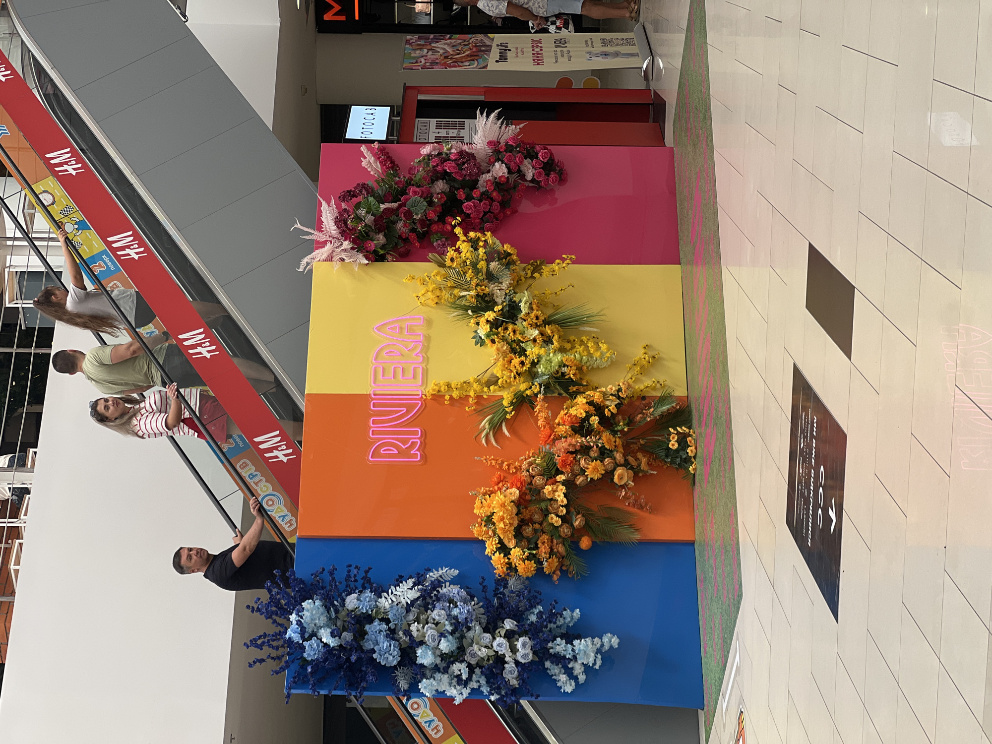 The image depicts an indoor shopping mall environment, possibly in Fontanka, Ukraine, as suggested. In the foreground, a large, colorful display with vibrant floral arrangements spans across panels of blue, orange, yellow, and pink, with the word "RIVIERA" illuminated in neon pink text on the yellow panel. The floor is tiled in a light color, with several dark floor decals, one of which displays text in a foreign language and an arrow pointing to the left.
In the midground, an escalator is visible, with several individuals ascending. A man in a black t-shirt is holding onto the escalator railing, looking towards the camera. Other individuals, including two women and a child, are also on the escalator. The red escalator railing is decorated with alternating logos of "H&M" and other text.
In the background, parts of a store are visible, including signage for "FOTOCAB" and other advertisements. The overall impression is of a public commercial space, likely a mall, with decorative elements and shoppers.