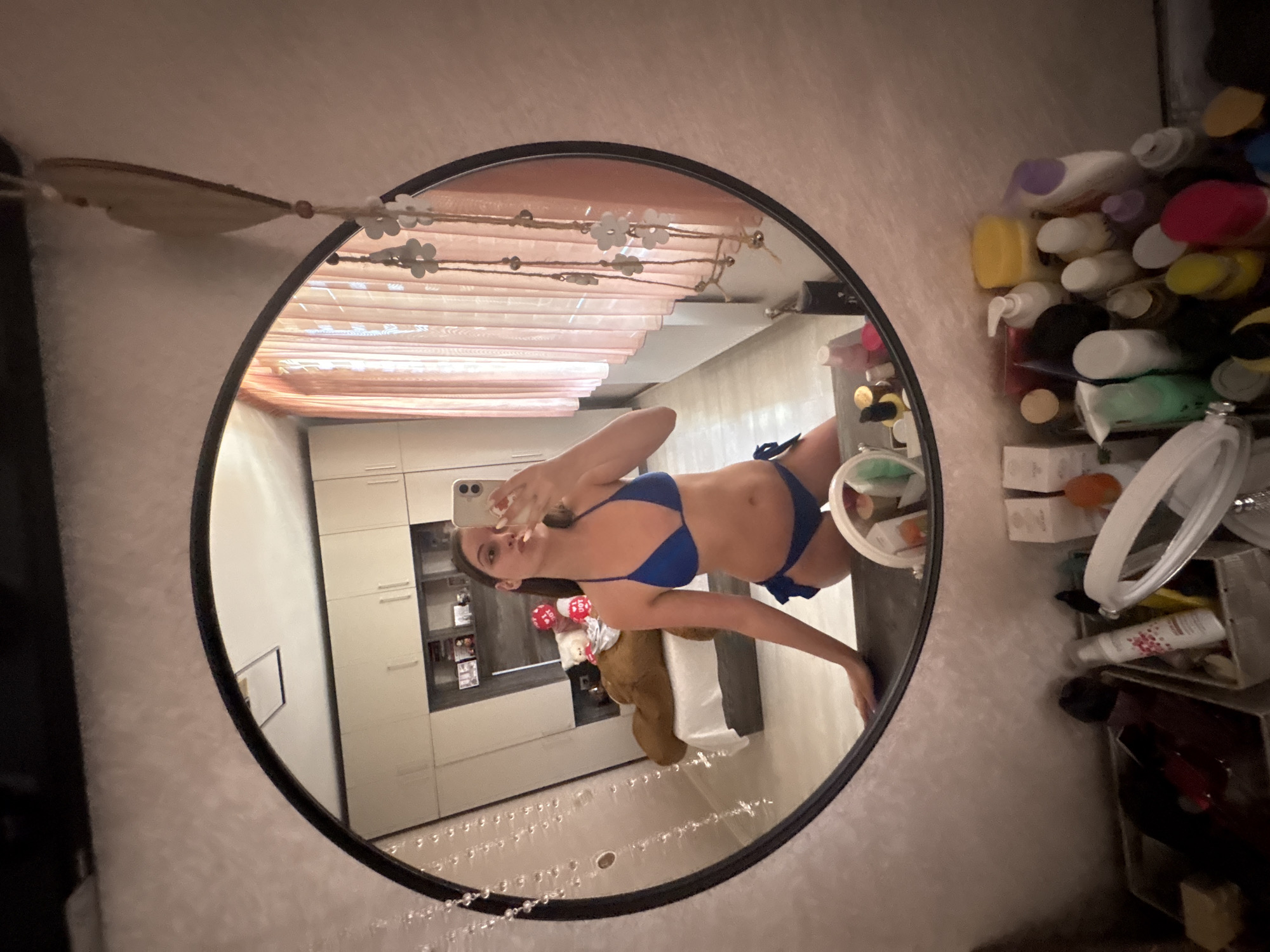 A young woman is captured in a mirror selfie, wearing a blue bikini. She is standing in what appears to be a bedroom, with a large teddy bear and a white duvet on the bed behind her. A tall, built-in wardrobe with cream-colored doors is visible to the left, and a set of pink curtains hangs on the right. The room is illuminated by soft lighting, and the overall atmosphere is casual and personal. The image was taken in Bulgarovo, Bulgaria.