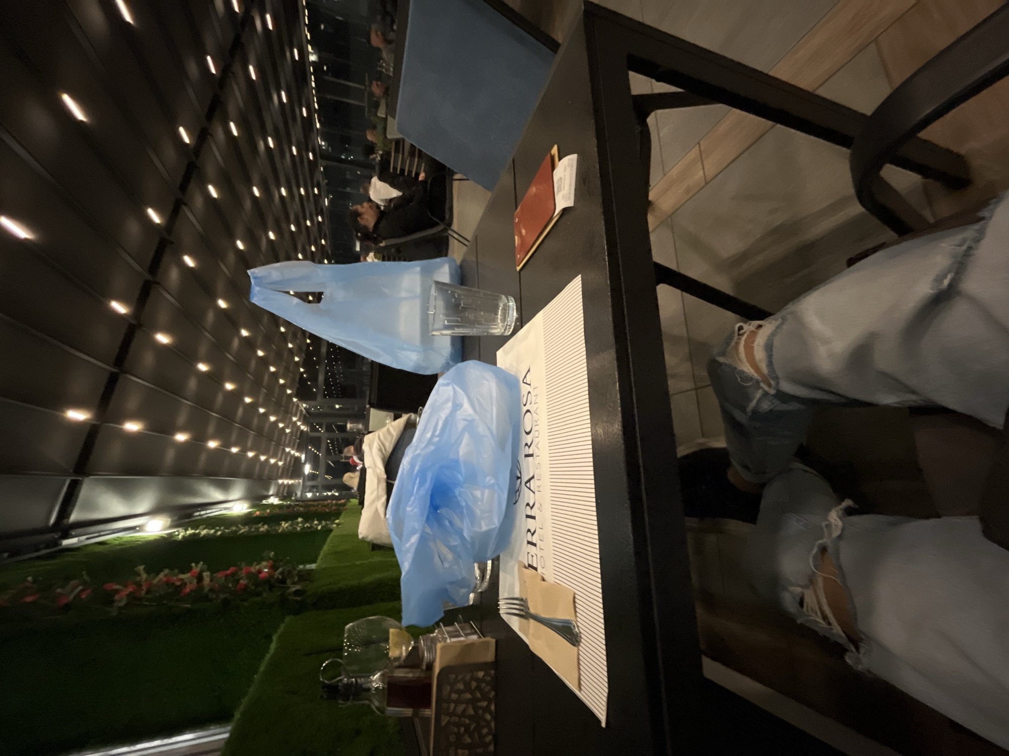 A tilted, low-angle shot depicts a table at Terra Rosa, a hotel and restaurant in Karlovo, Bulgaria. The table is set with a menu, silverware, a water glass, and two blue plastic bags. Outside the establishment is a wall of lights, greenery, and flowers. An individual wearing ripped jeans is seated at the table. Other patrons can be seen in the background.