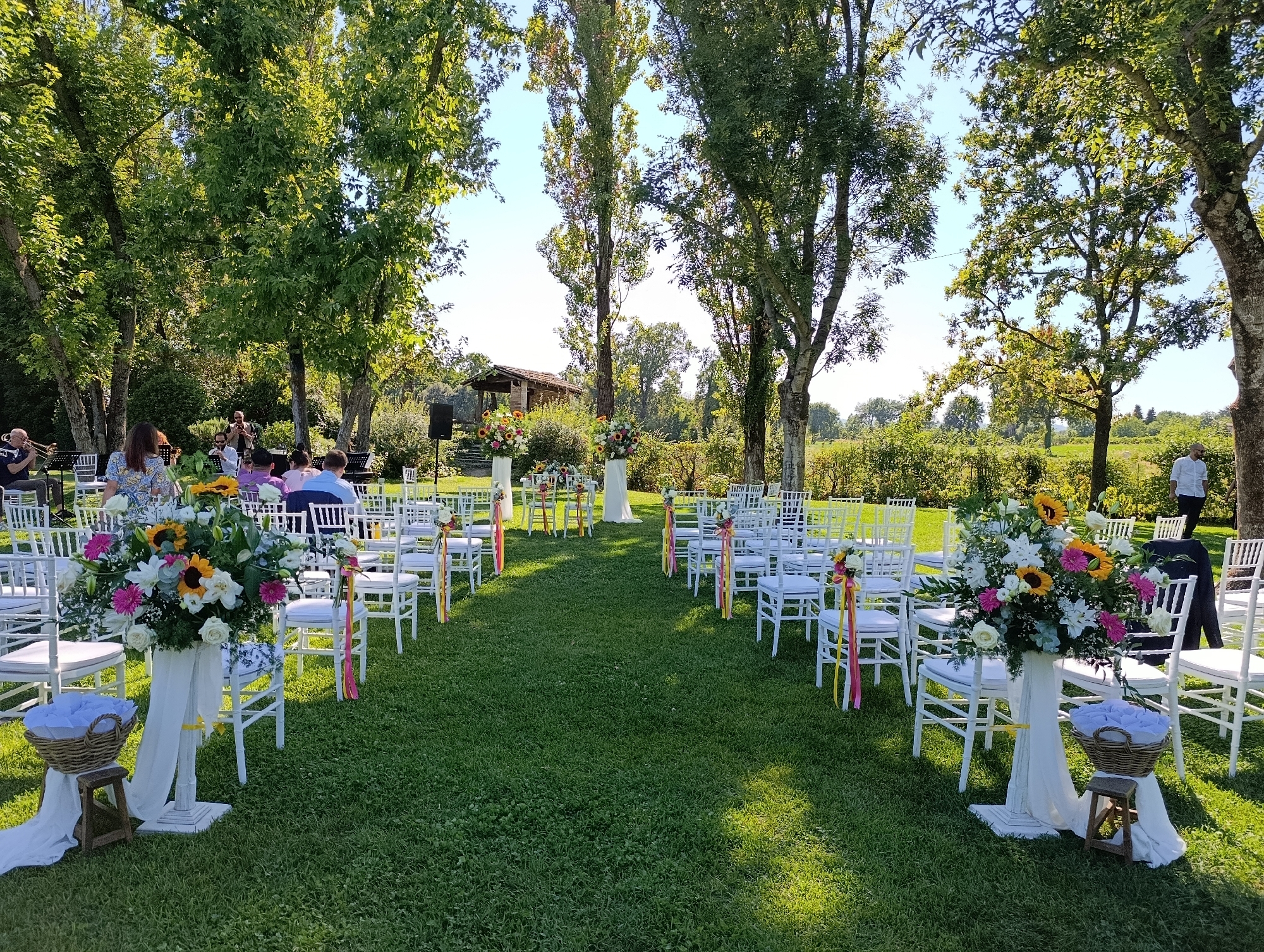 The image depicts an outdoor wedding ceremony setup in a grassy field surrounded by trees and vegetation. Two rows of white chairs are arranged facing a central aisle, with floral arrangements decorating the aisle and some chairs. In the background, there are trees, a building with a rustic roof, and a clear sky. Several people are visible, some seated in chairs and others standing, appearing to be guests or participants in the event. One person is visible in the far right, wearing a white shirt. The scene is set on a sunny day, with shadows cast on the grass, suggesting it is afternoon. The overall ambiance is festive and organized, consistent with a wedding ceremony. The provided information states the location is in the city of Scandiano, Italy.