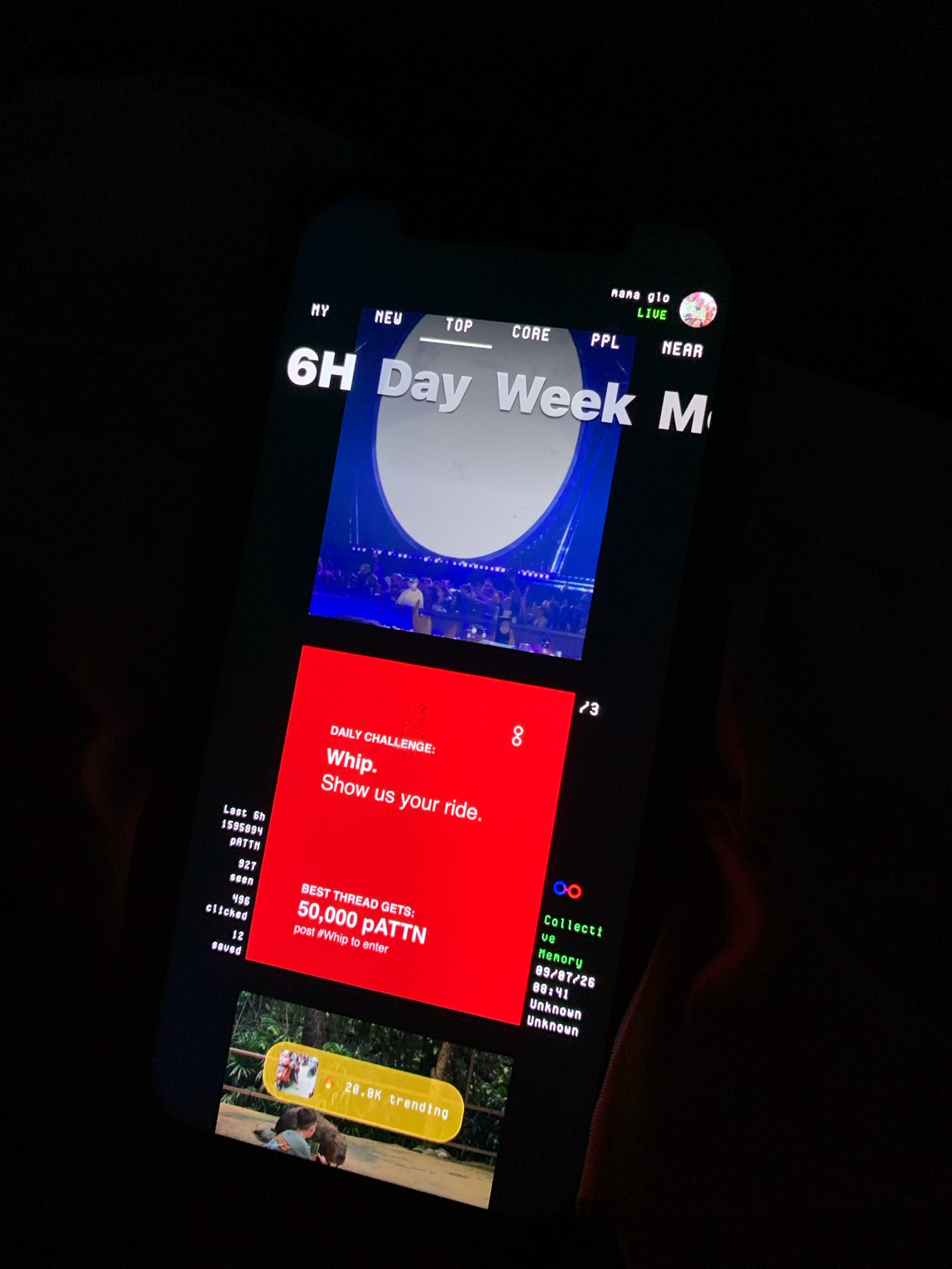 The image shows a close-up of a smartphone screen displaying a social media app interface. The screen is dark, with bright text and graphics visible. The app appears to be a feed or timeline, with various posts and sections.
At the top, there are navigation tabs labeled "MY," "NEW," "TOP," "CORE," "PPL," and "NEAR." Below these tabs, large white text reads "6H Day Week M." Further down, there is a video or image displayed within a blue frame, showing a crowd of people at an event.
To the left of the main content, there's a vertical list of metrics: "Last 6h," "1595894," "PATTH," "927," "seen," "486," "clicked," and "12 saved."
A prominent red section on the right contains a "DAILY CHALLENGE: Whip. Show us your ride." Below this, it states, "BEST THREAD GETS: 50,000 pATTN" with instructions to "post #Whip to enter."
At the bottom of the screen, a smaller image shows a person interacting with an animal, with a yellow banner displaying "20.0K trending." Another section on the right has the text "Collective Memory" with a date and time "09/07/26 08:41" followed by "Unknown."
The overall scene is dark, suggesting the photo was taken in low light conditions, possibly at night. The image is captured from an angle, and the phone is being held by a person whose hands are partially visible at the bottom and right edges of the frame. There is no discernible information about the city of Ado, Nigeria, in the provided image.