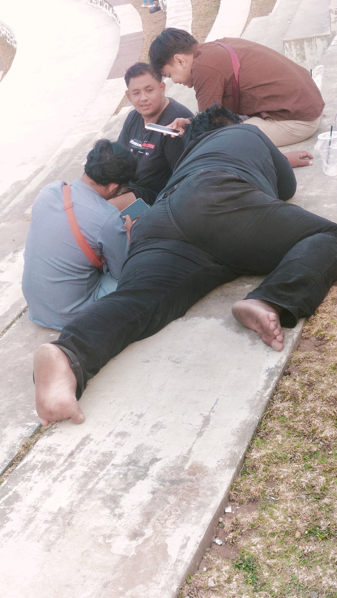 The image depicts four individuals in an outdoor, urban environment, likely in Ketanggan, Indonesia, given the context. Three men are seated on a concrete structure, possibly part of a park or public seating area with tiered levels.
In the foreground, a man is lying on his side, facing away from the camera. He is wearing black pants and is barefoot, with his feet positioned on the concrete. His right foot is visible, showing dirt.
To his left, a man in a light blue shirt with an orange strap across his shoulder is seated and looking down at a mobile device he is holding.
Behind these two, another man in a black t-shirt is seated and looking towards the man lying down, holding a white mobile device.
Further back and to the right, a man in a brown shirt and light-colored pants is also seated, leaning forward and looking down, seemingly at something on his lap or in his hands, obscured from view.
The environment appears to be a public space with concrete walkways and grassy areas. The lighting suggests it is daytime. A plastic cup with a straw is visible on the concrete to the right of the man in the brown shirt. The overall scene suggests a casual gathering or break in a public area.
