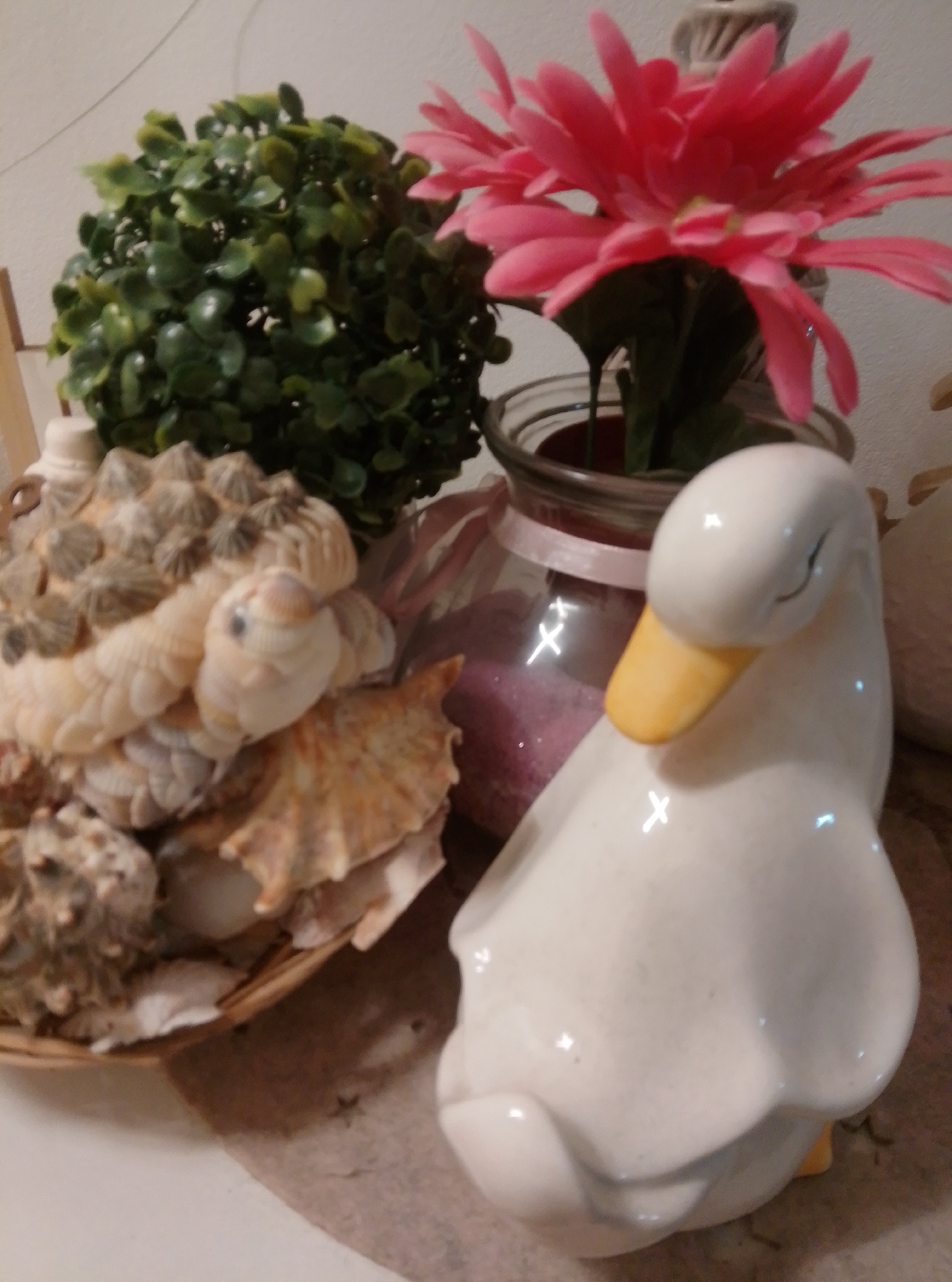 A white ceramic duck figurine with a yellow beak is positioned in the foreground, on the right side of the image. Behind the duck, a glass jar filled with pink sand is visible, tied with a pink ribbon. A bright pink artificial flower with multiple petals is placed inside the jar, with its stem extending upwards. To the left of the jar is a round, artificial green bush. In front of the bush and jar, a collection of seashells is arranged in a basket. The shells are a mix of cream and brown colors, with some appearing to be barnacles. The scene is set on a light-colored surface, possibly a table, with a wall visible in the background.