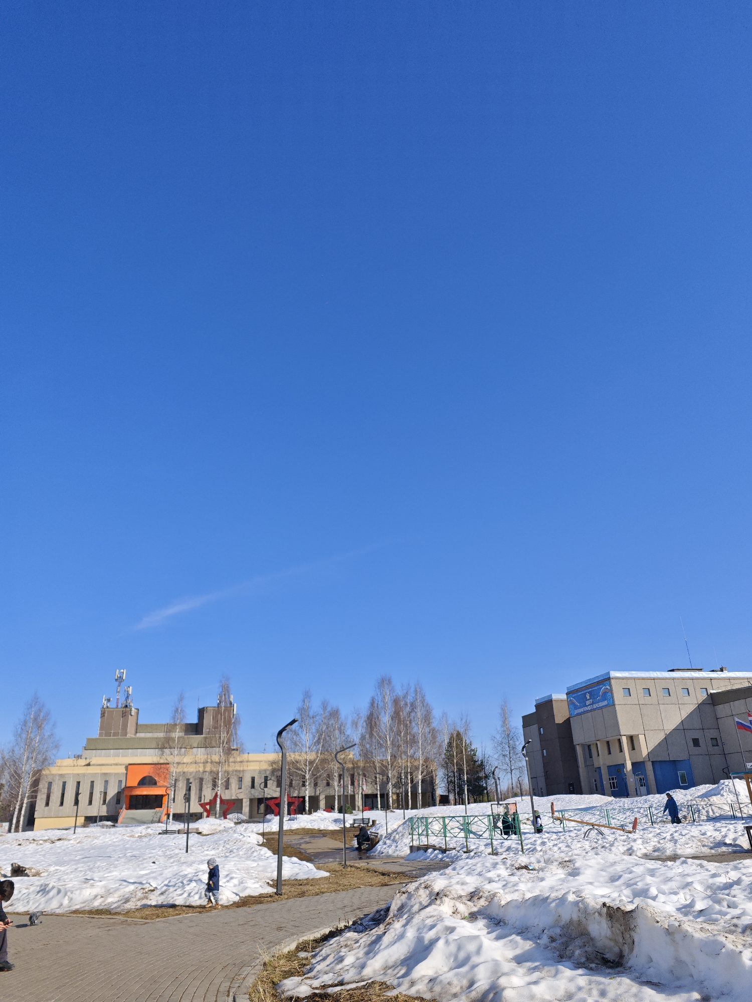 The image depicts a winter scene in Марковский, Russia. Patches of snow cover the ground in a park or public space. A paved walkway meanders through the snow-covered areas. In the background, two buildings are visible. The building on the left has a distinctive architectural design with multiple tiers and visible antennae on the roof, and features orange coloring around the entrance. To the right of this building, there's a row of birch trees. The building on the right is a gray, rectangular structure with a visible mural on one side. Between the buildings is a small playground area with play equipment. Several people can be seen in the vicinity, including a child walking along the path, individuals on the playground equipment, and someone standing near the buildings. The sky is clear and blue.